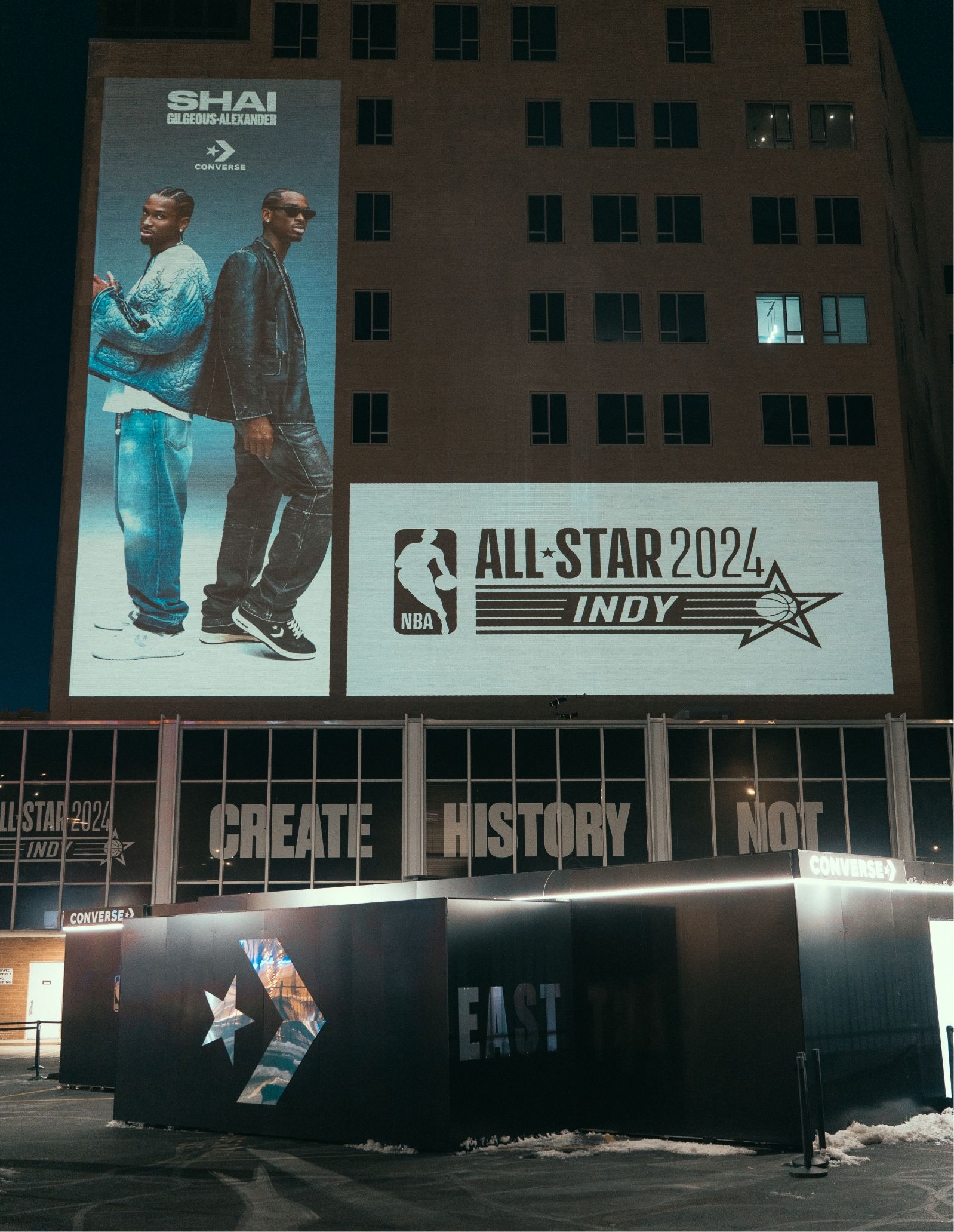 Night time shot of 1 story black building with mirrored converse logo in foreground. Background shows a large building with two billboards. One is Converse Choose your weapon with Shai the other is NBA All Star 2024 Indy.