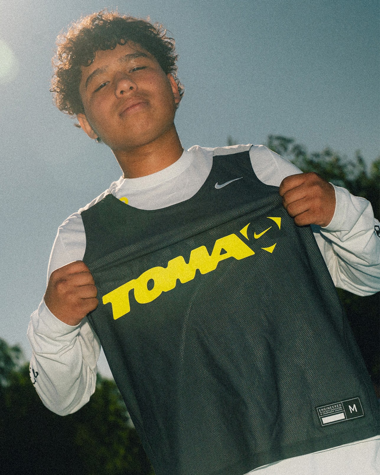 A young person stands outdoors holding up a dark sports jersey with the word “TOMA” in bold yellow letters. The sun is shining in the background.