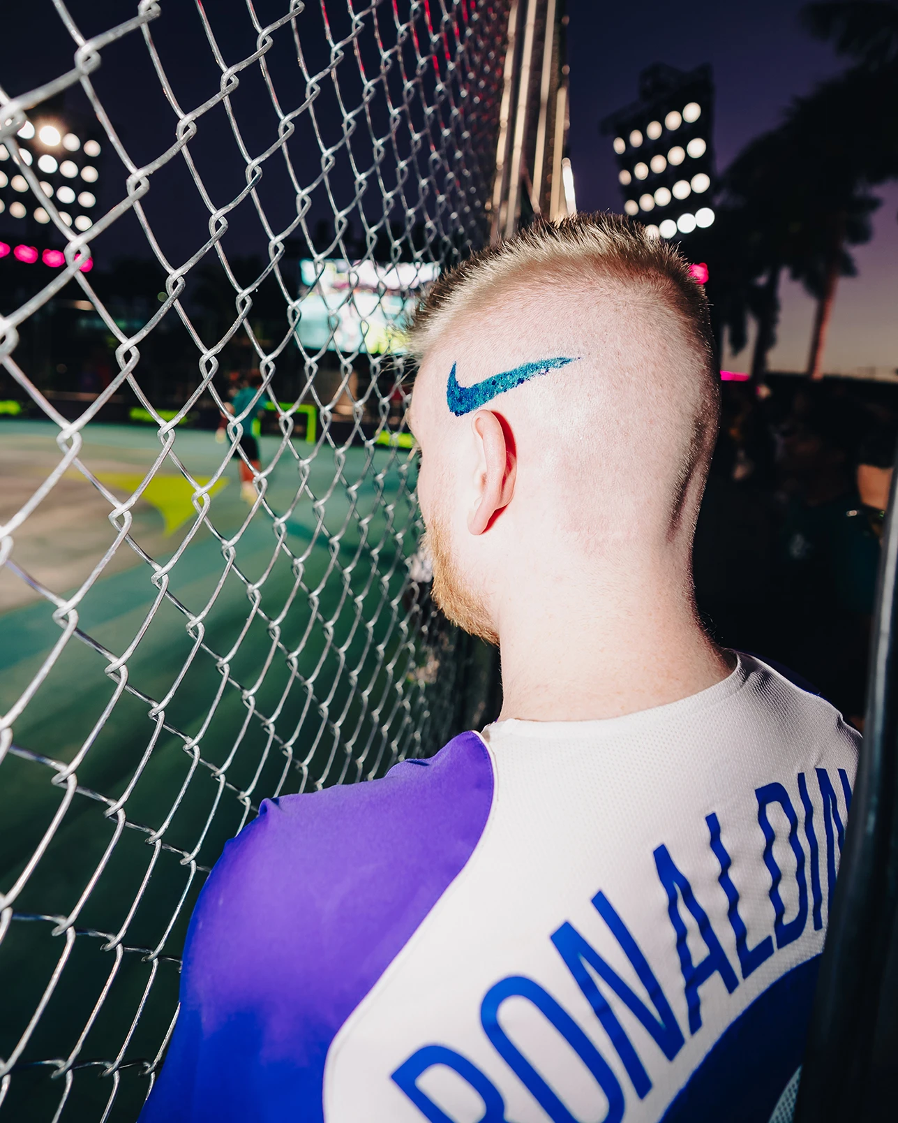A person with a blue Nike swoosh shaved into their hair wears a jersey with “RONALDINHO” on the back, standing by a chain-link fence at a lit outdoor sports venue.