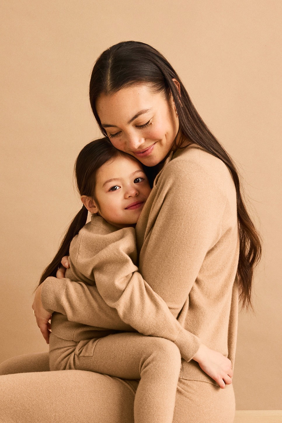 A woman sits and hugs a young girl, both wearing matching beige outfits, against a beige background.