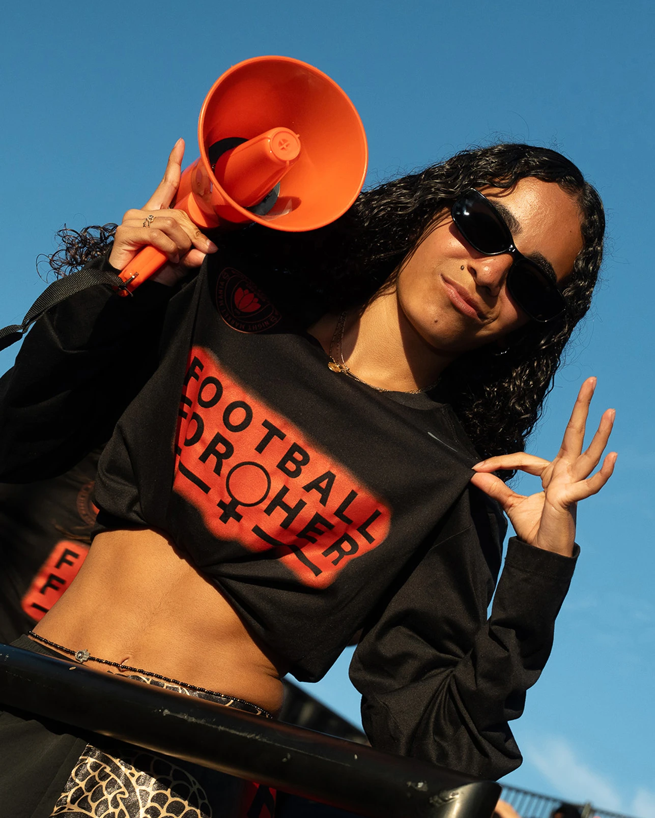 A person in sunglasses holds an orange megaphone and makes an “OK” sign, wearing a black crop top reading “FOOTBALL CROHER” against a clear blue sky.