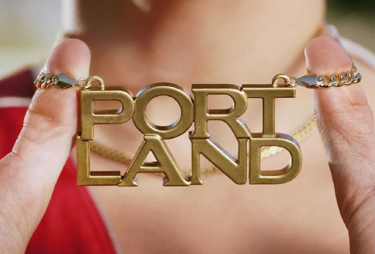 A person holds a gold necklace with large letters spelling "PORTLAND" in two lines, close to their neck.