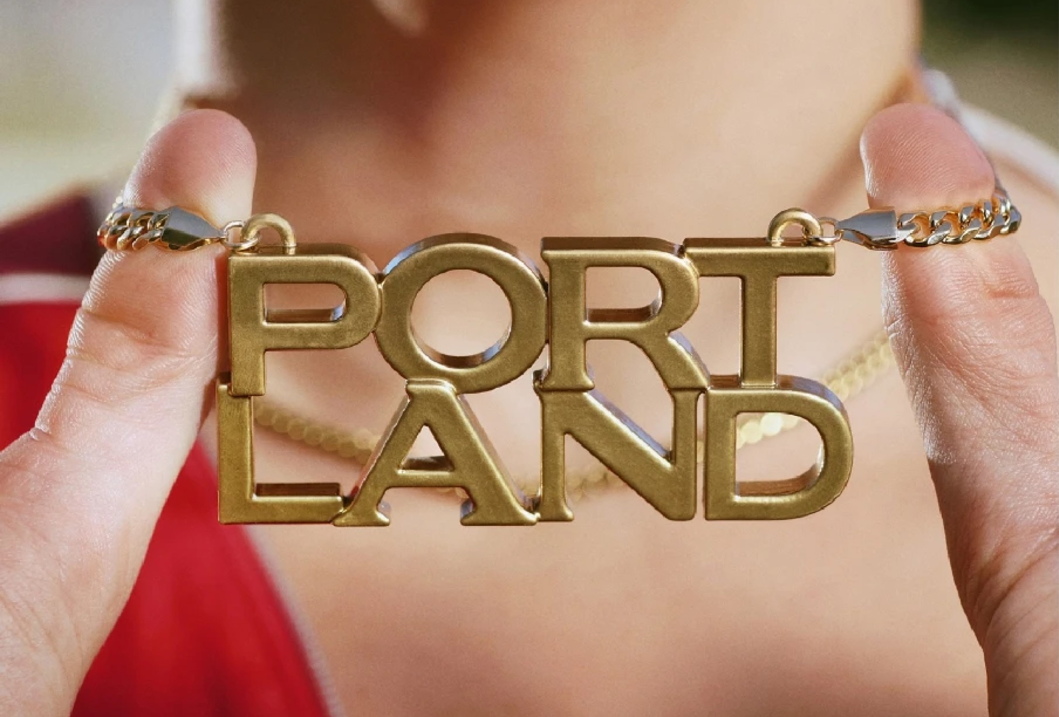 A person holds a gold necklace with large letters spelling "PORTLAND" in two lines, close to their neck.