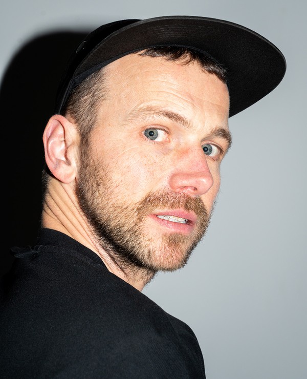 A man with short hair, light eyes, and facial stubble wearing a black cap and shirt looks over his shoulder against a plain background.