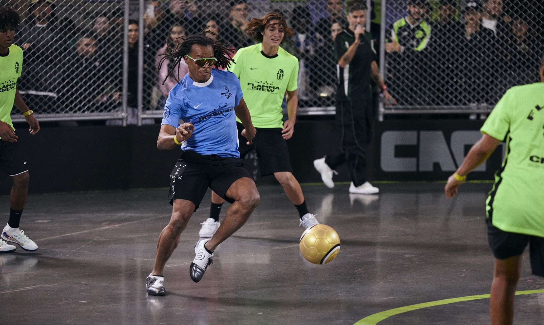 A football player wearing a blue jersey and sunglasses kicks a gold ball on an indoor court during a match. Spectators watch from behind a chain-link fence.