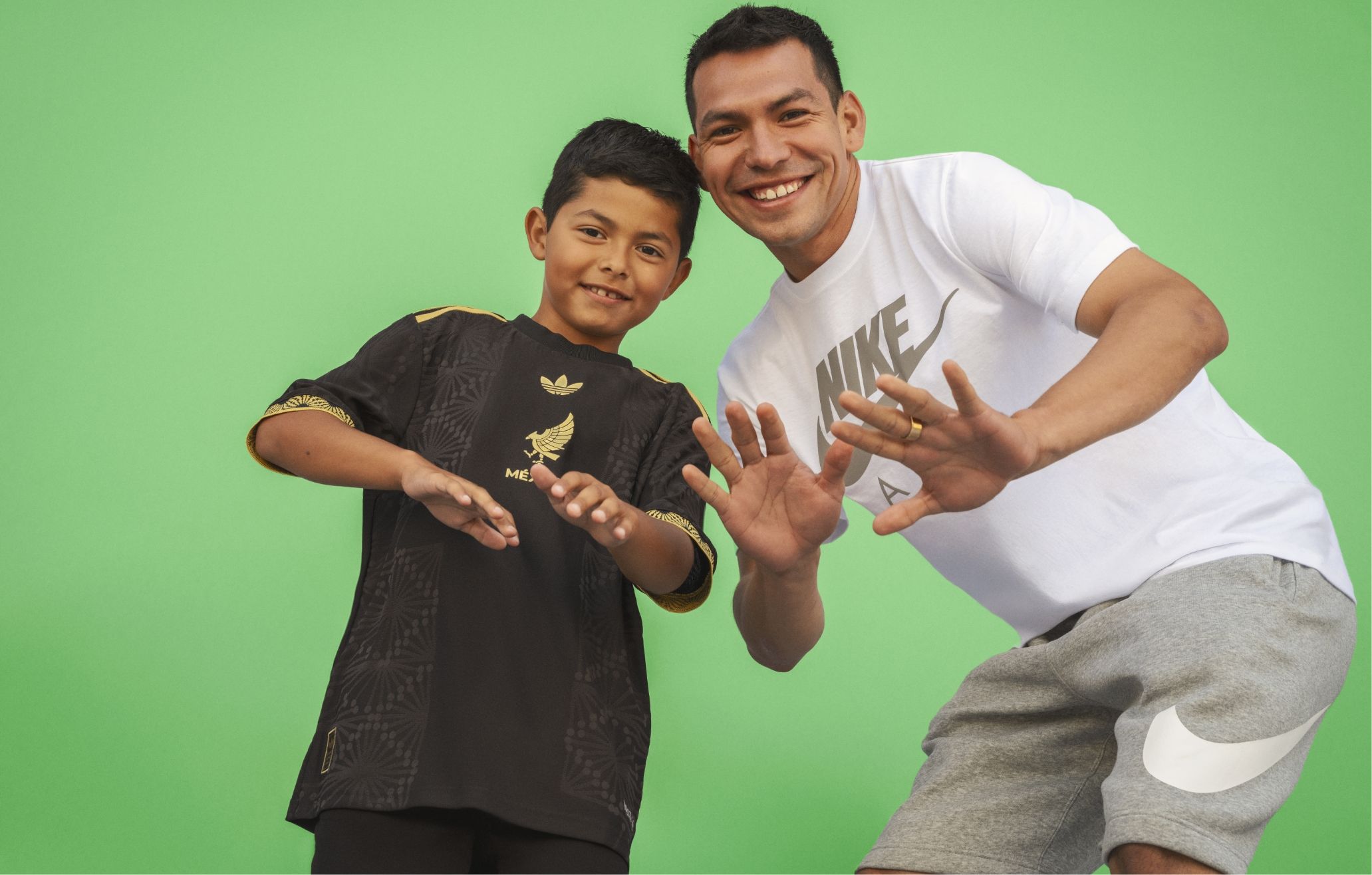 An adult and a child smiling and posing with hands up in front of a green background. The child wears a black sports jersey, and the adult wears a white Nike shirt and gray shorts.