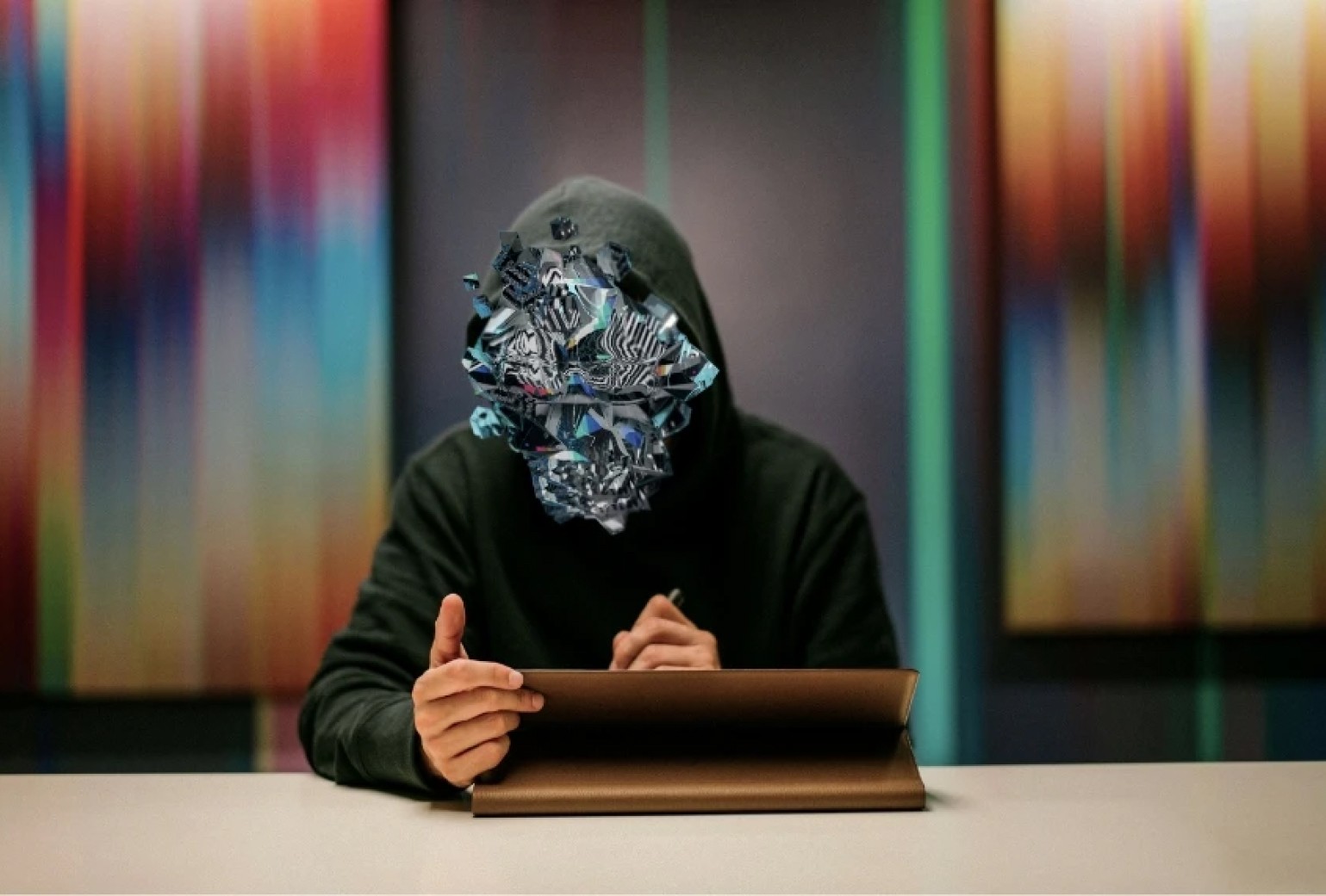 A person in a hooded sweatshirt sits at a desk using a tablet, with their face replaced by a digital, abstract geometric design.