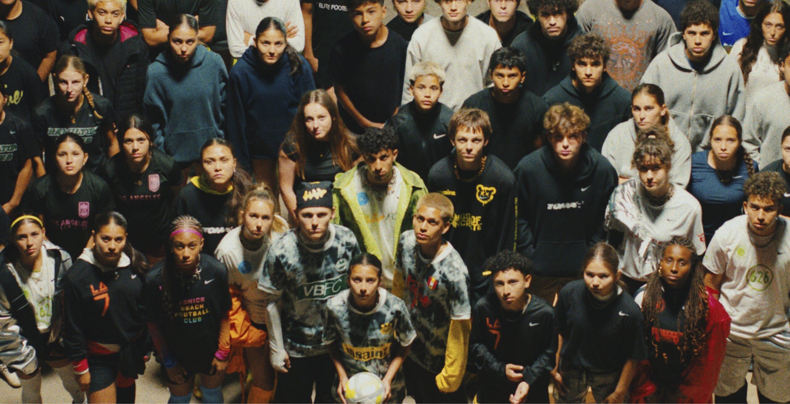 A large group of young people wearing casual clothing stand close together, looking up at the camera with serious expressions.