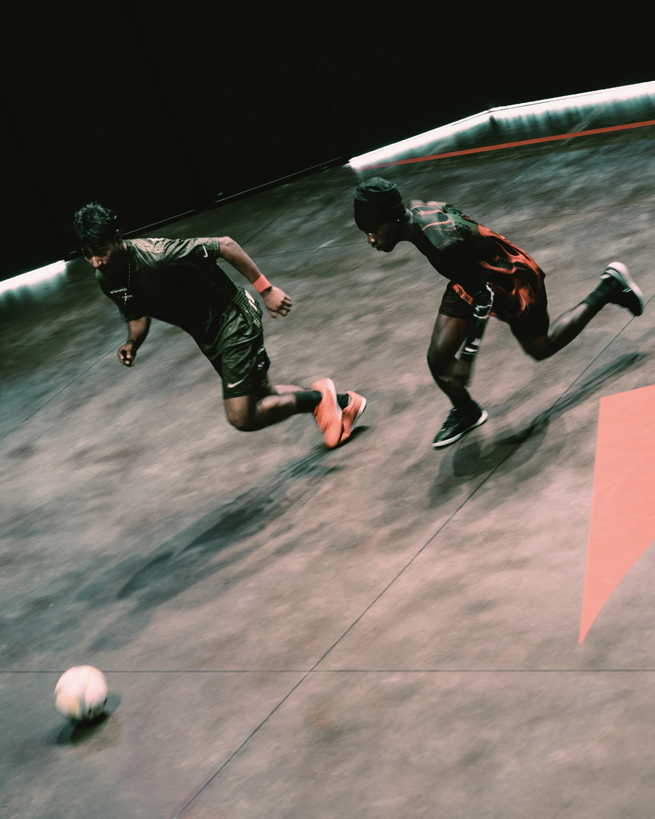 Two people in sportswear running after a soccer ball on an indoor court with a dark background and red lines on the floor.