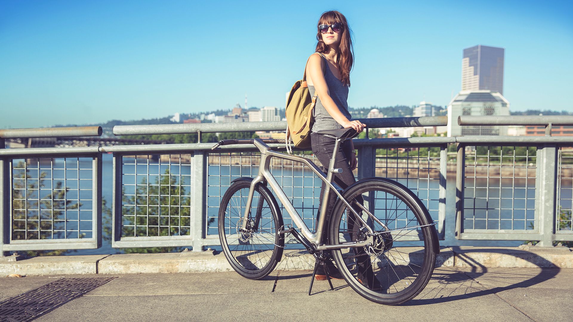 Woman in sunglasses and backpack standing with her bike on a city bridge, overlooking a river and skyline on a sunny day.