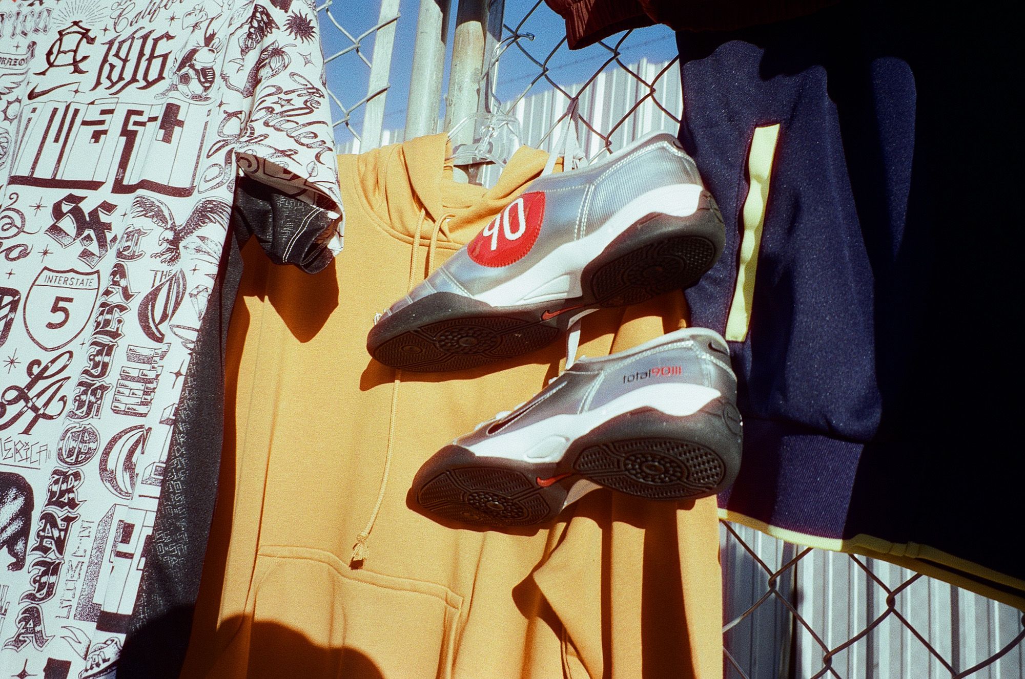 A yellow hoodie, a graphic T-shirt, and a pair of gray sneakers with red detailing hang on a fence in sunlight.