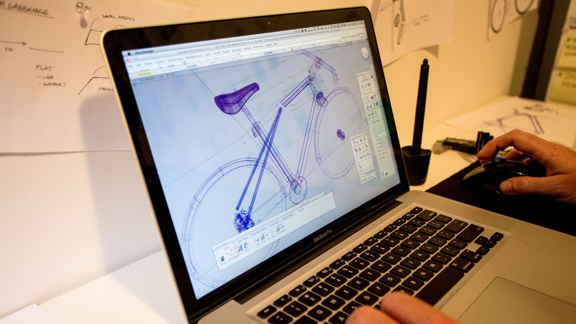 A laptop screen displays a cad design of a bicycle, with a person's hand using a mouse on the desk, surrounded by papers and pens.