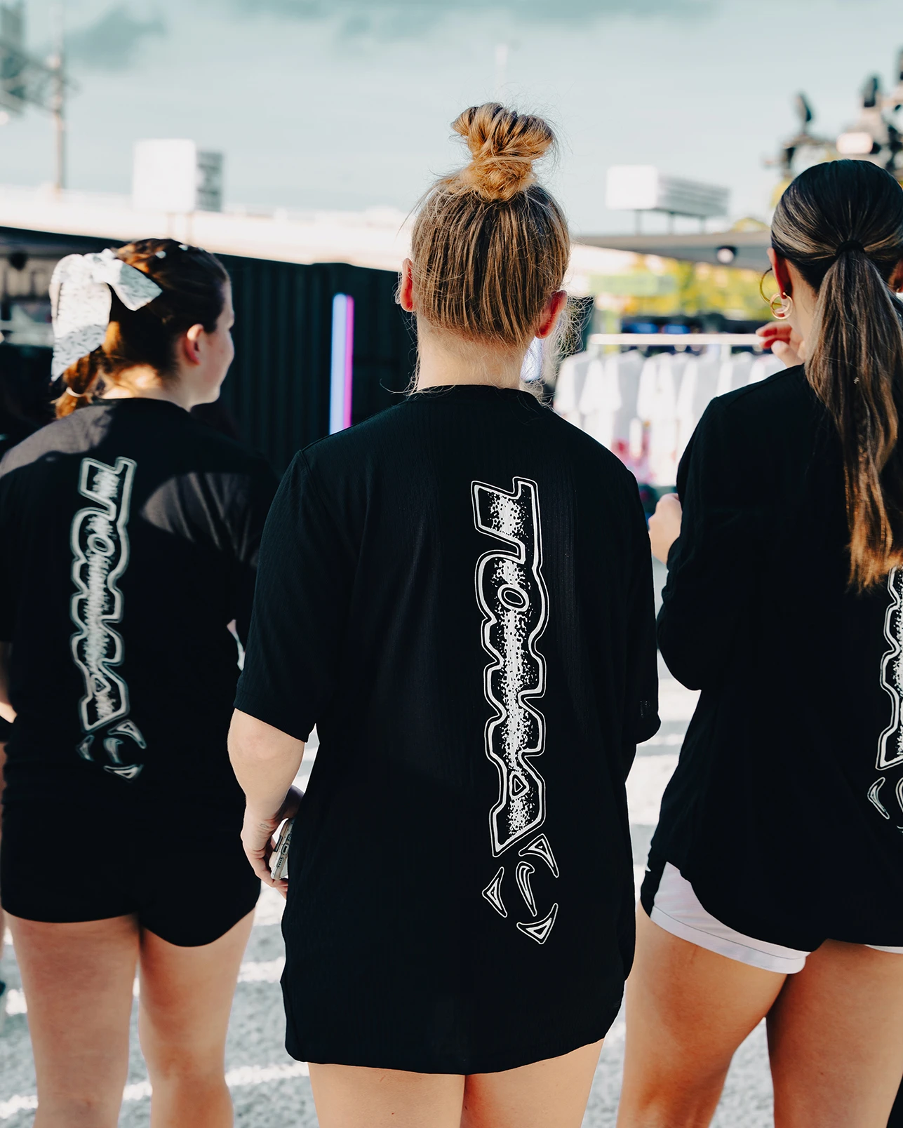 Three people wearing black shirts with white graphic text on the back stand outdoors, facing away from the camera on a sunny day.