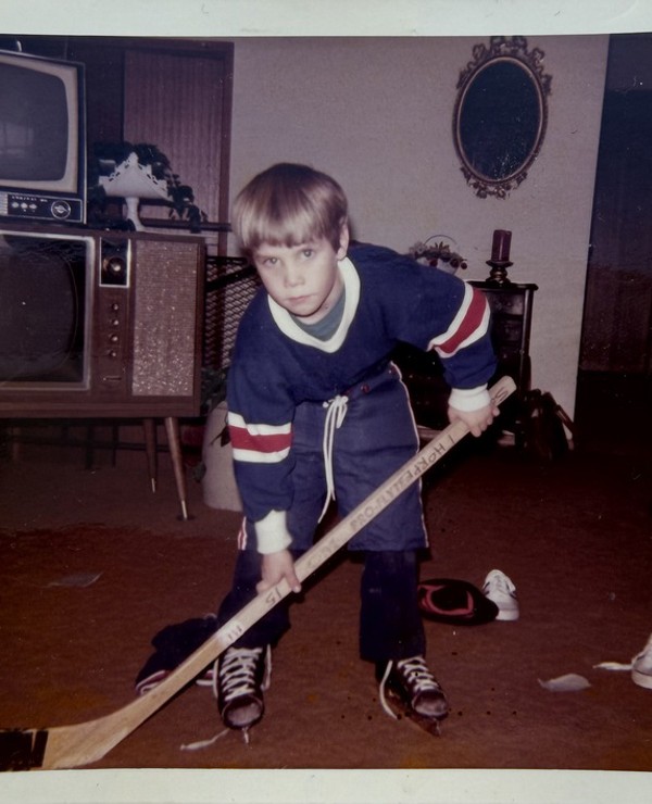 Young boy in a hockey uniform poses with a hockey stick indoors on a carpeted floor, with vintage televisions and furniture in the background.