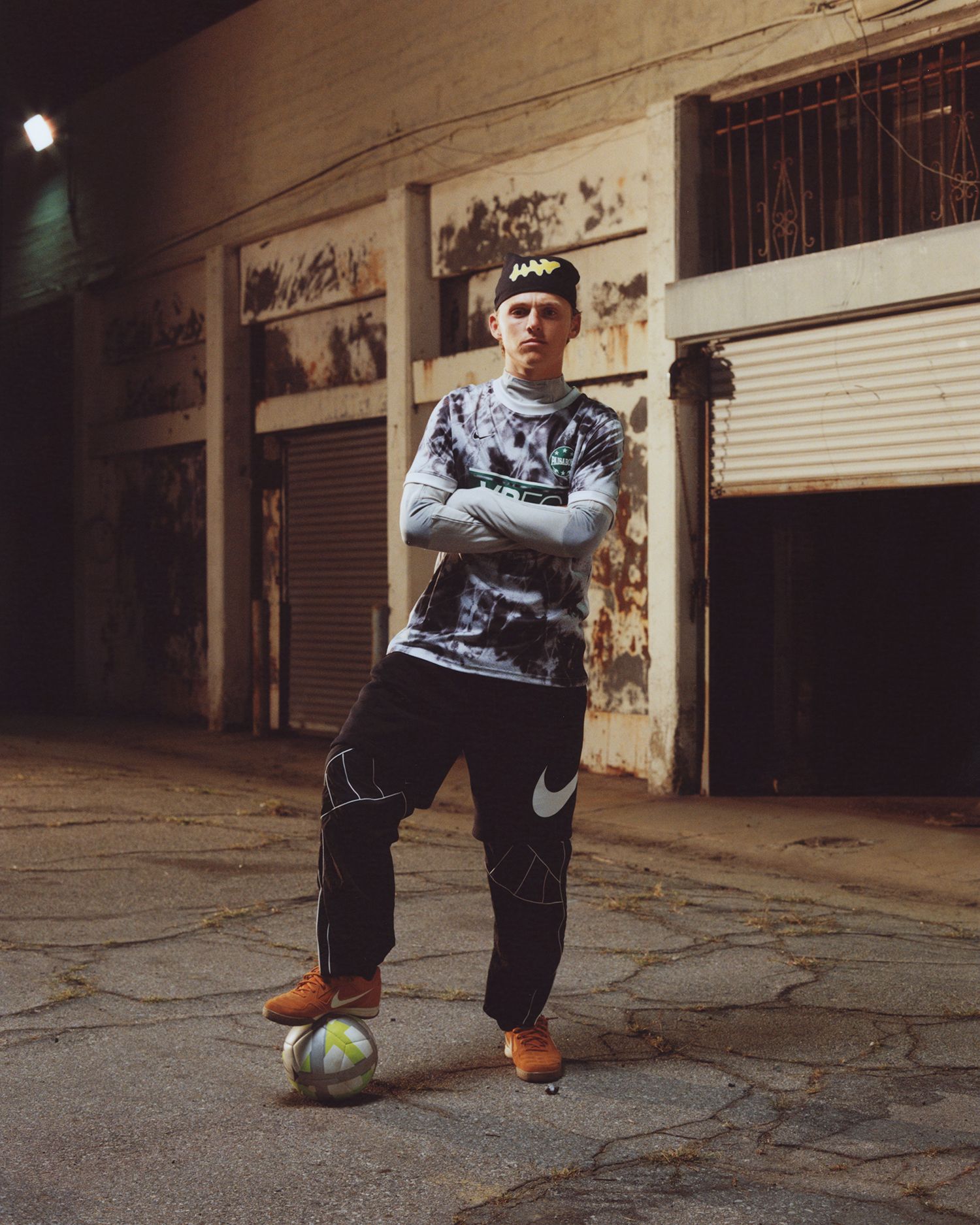 A person in sportswear stands on a cracked pavement at night with one foot on a soccer ball, in front of a worn building with closed metal shutters.