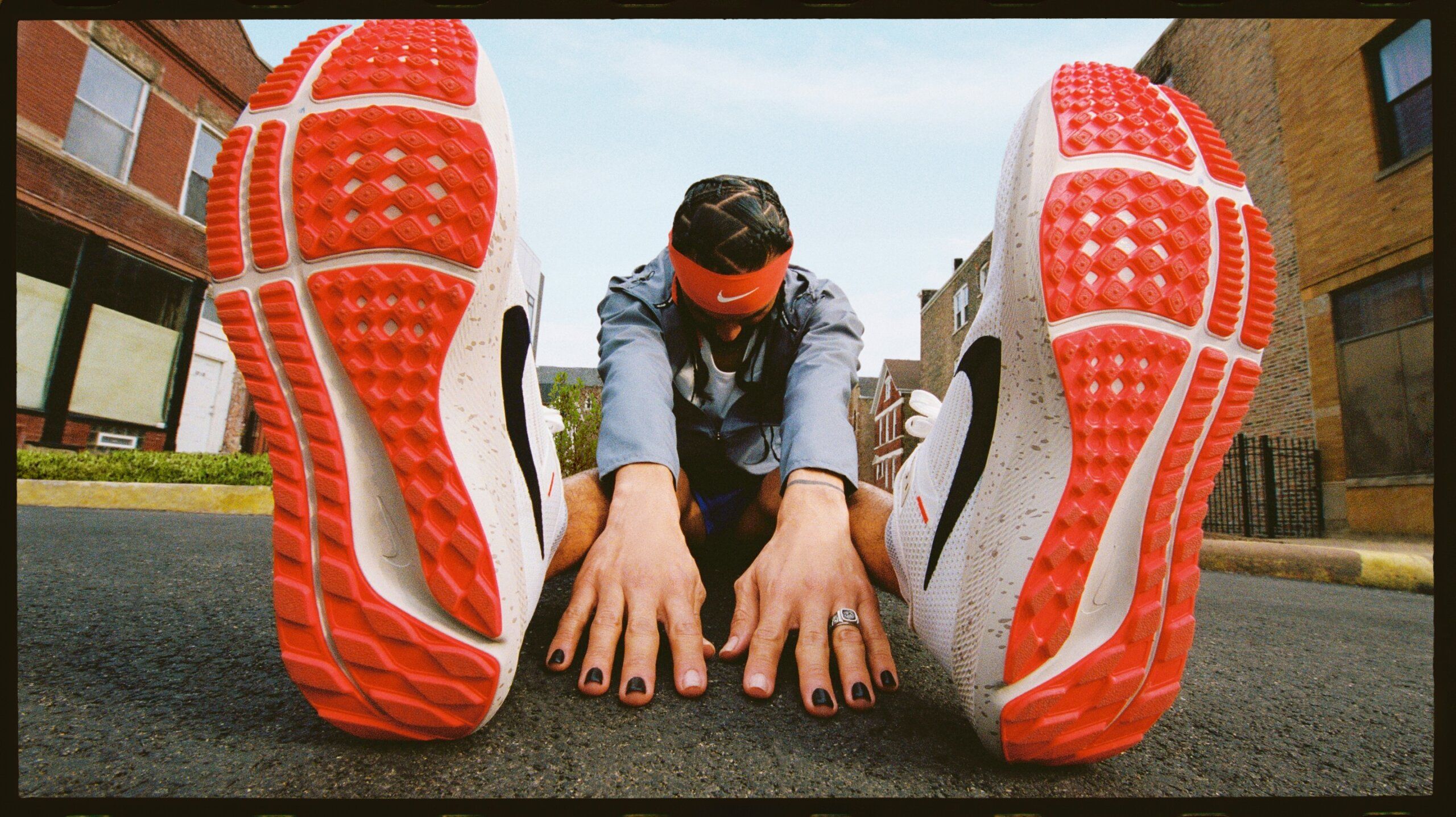 Man sitting in street stretching in seated forward bend. Bright red soles of Nike pegasus are prominent. Man has nails painted black and orange nike head band on