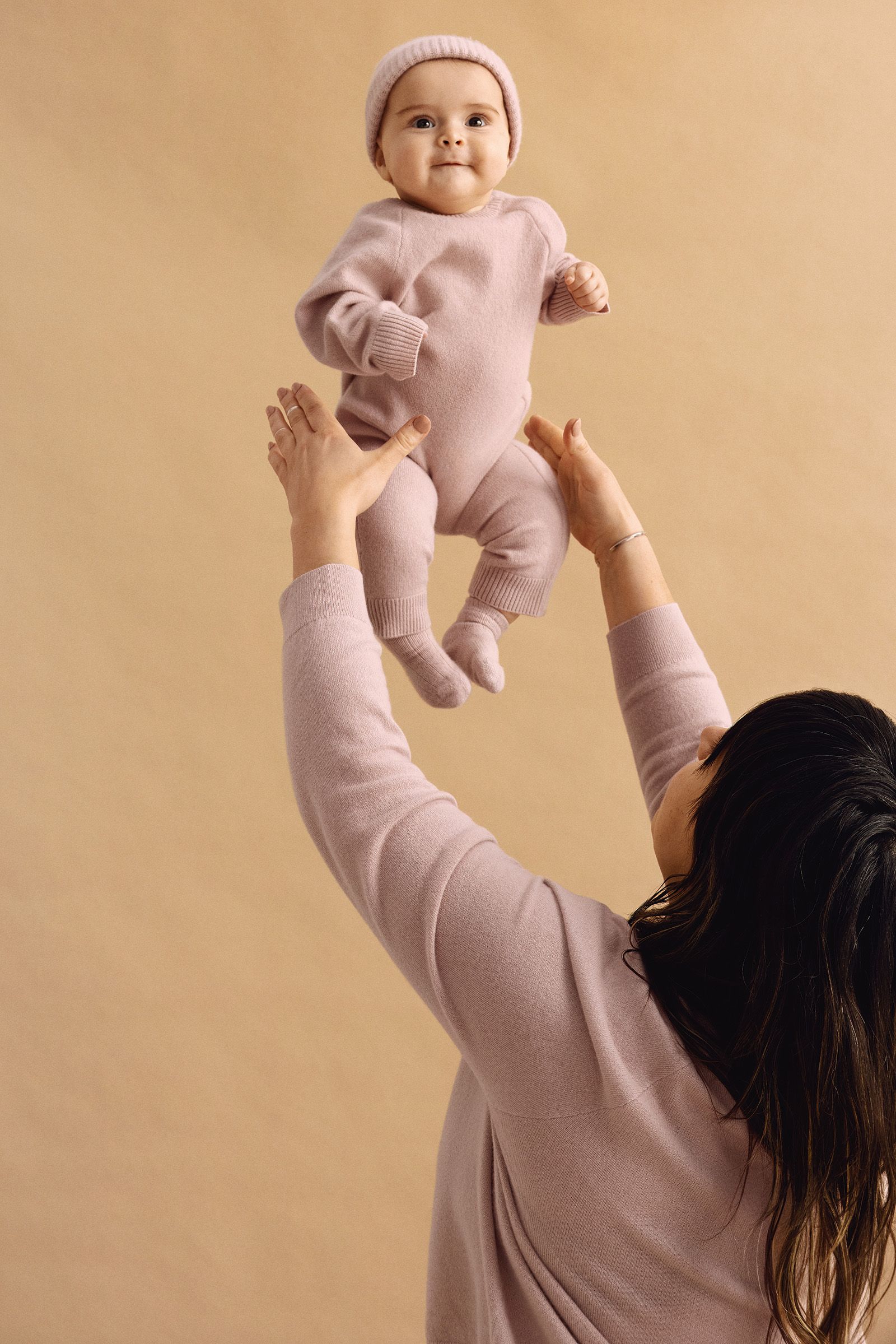 An adult lifts a smiling baby dressed in a matching pink knit outfit and hat against a beige background.