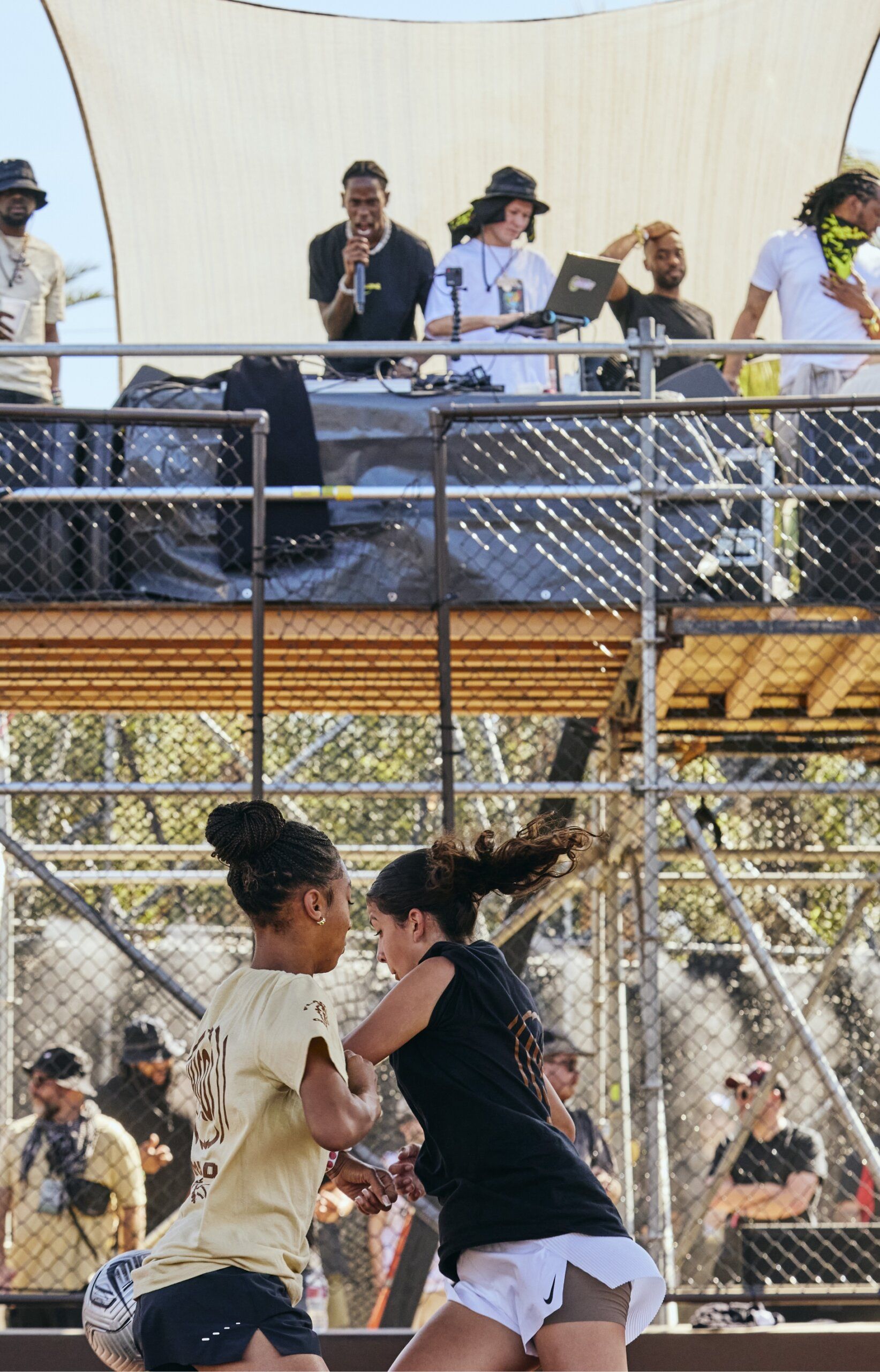 Two women compete for a basketball while a group of people, including a DJ, watch from an elevated platform behind a chain-link fence.