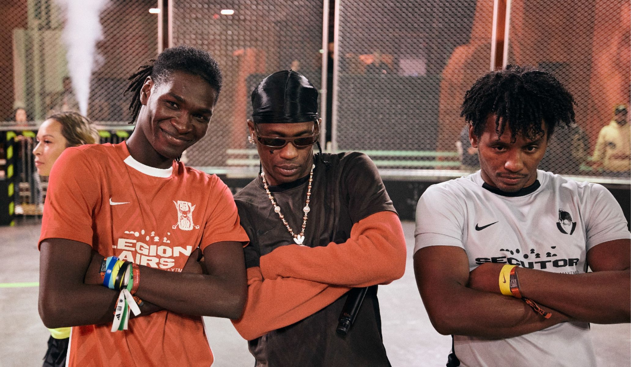 Three people including Travis Scott pose confidently with arms crossed inside a sports venue, wearing casual athletic clothing.