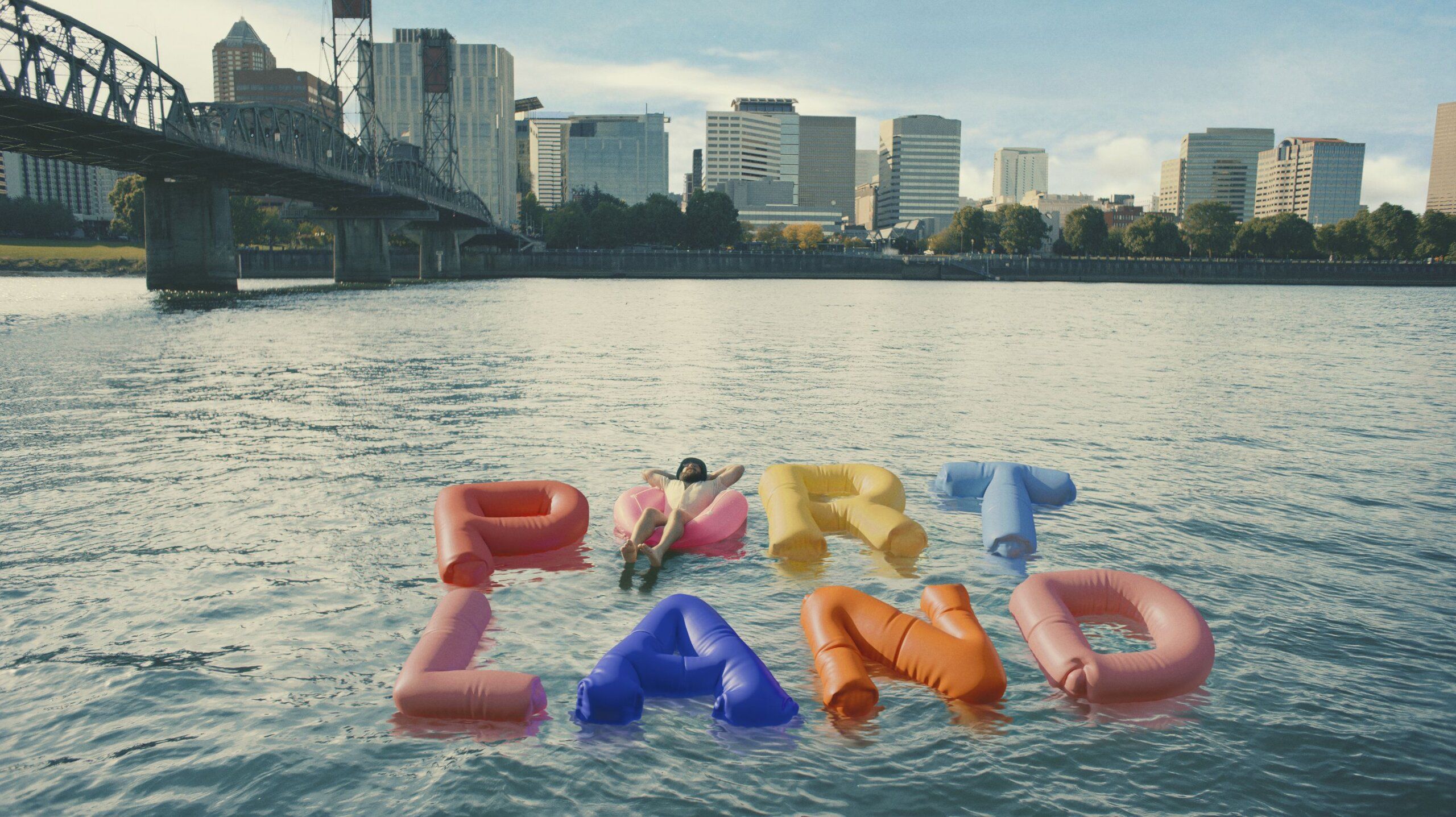 A person floats on a river among large inflatable letters spelling "PORTLAND," with a city skyline and bridge visible in the background.