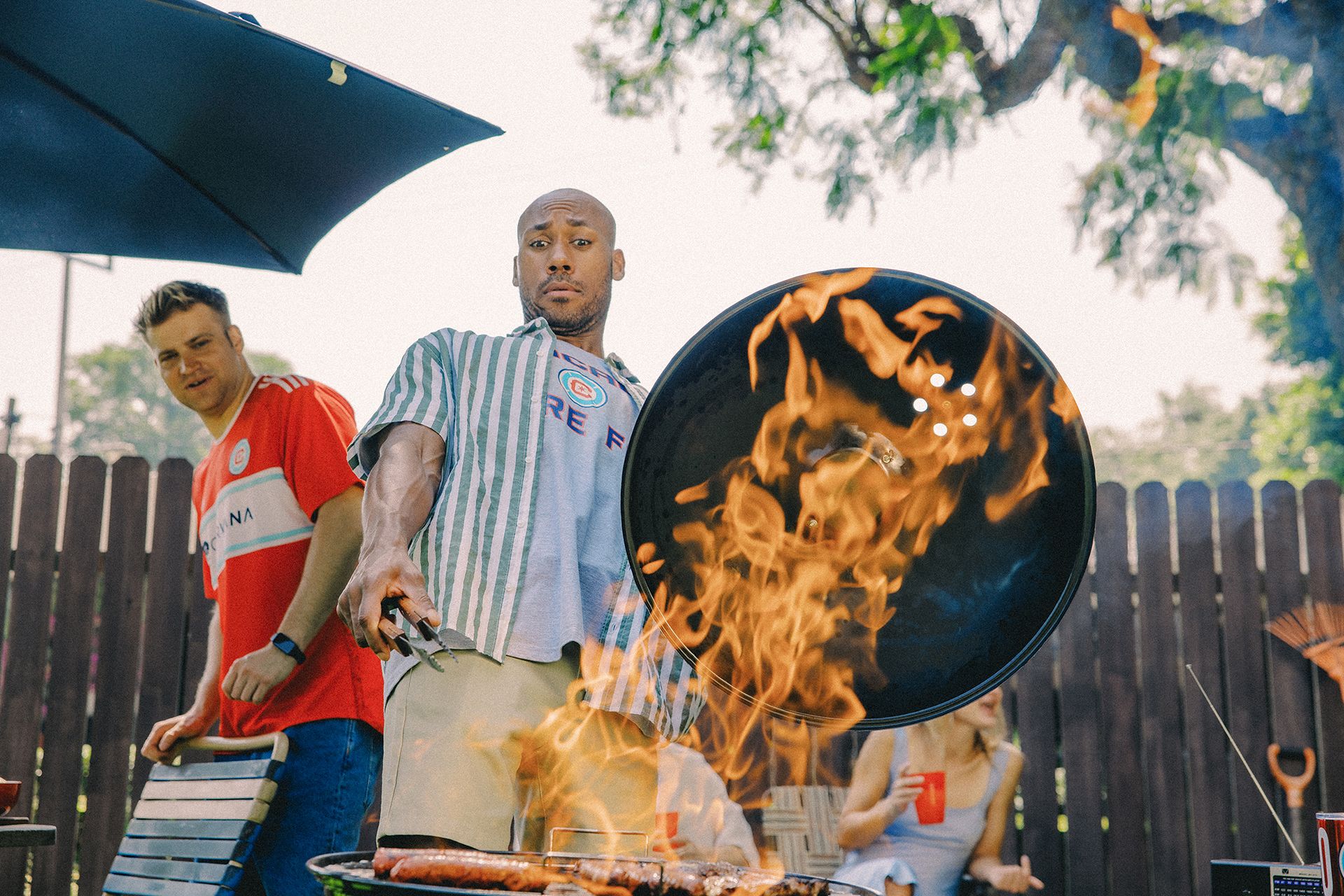 A man grilling food over high flames, with people around him. One person looks back while holding utensils, and another sits in the background holding a red cup.