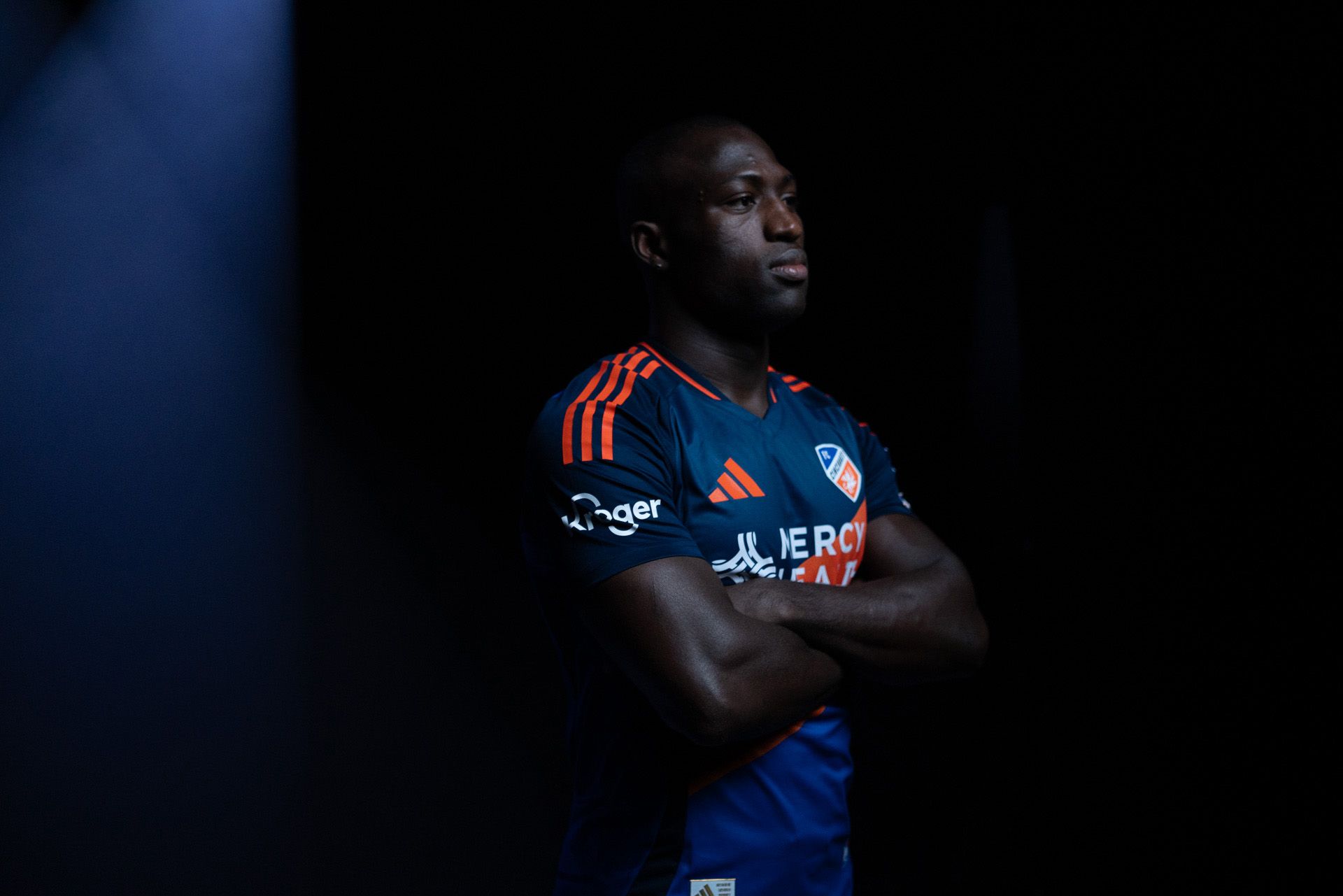 A male soccer player stands with arms crossed, wearing a dark blue jersey with orange stripes and logos, set against a dark background.