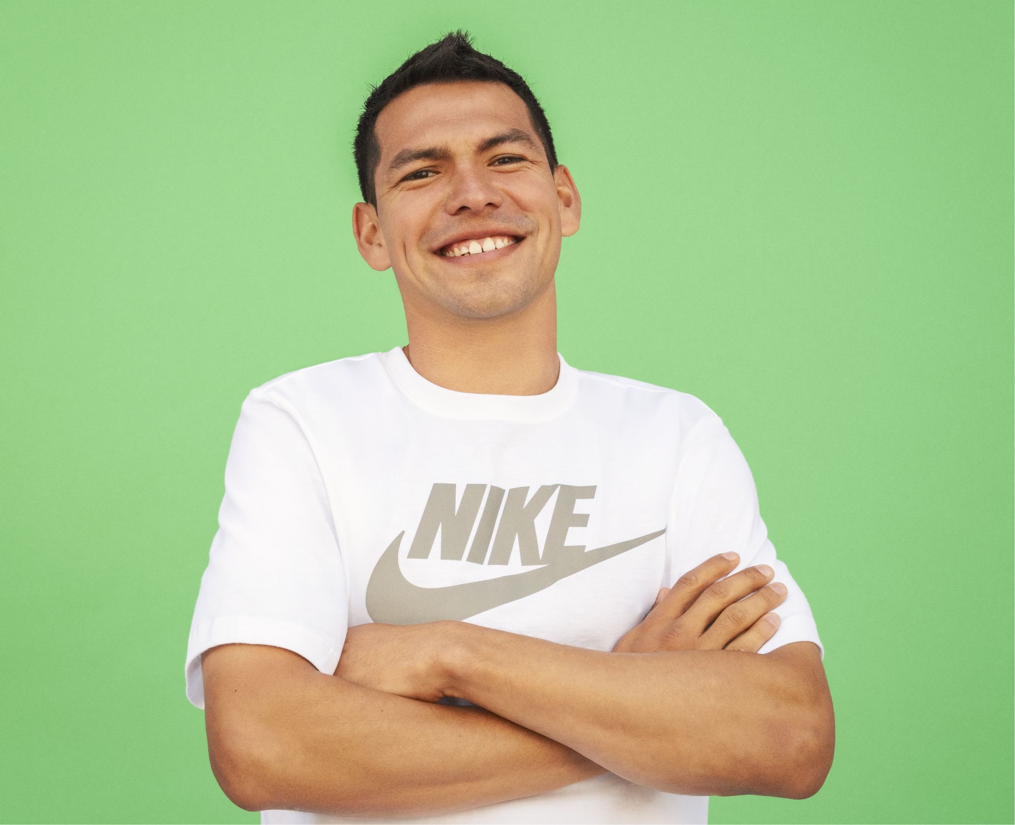 A man in a white Nike t-shirt stands with his arms crossed, smiling, in front of a plain green background.