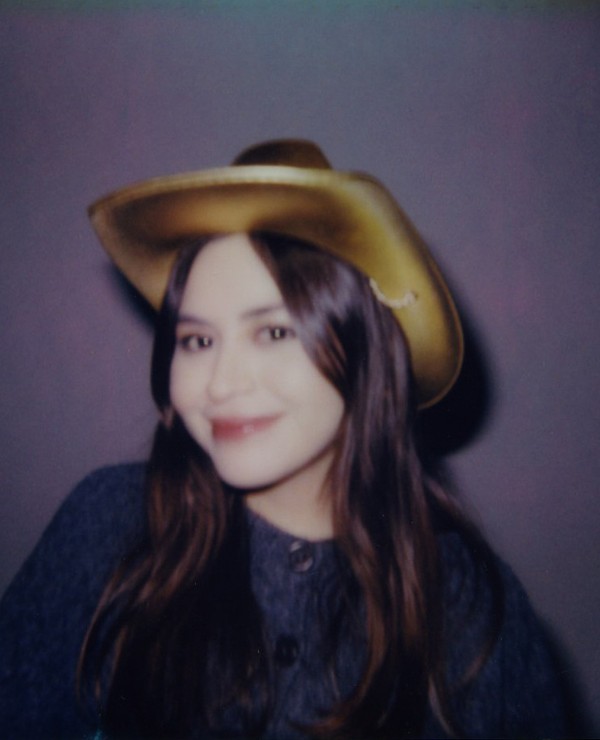 A person with long brown hair, wearing a gold cowboy hat and a dark buttoned shirt, smiles at the camera against a plain background.