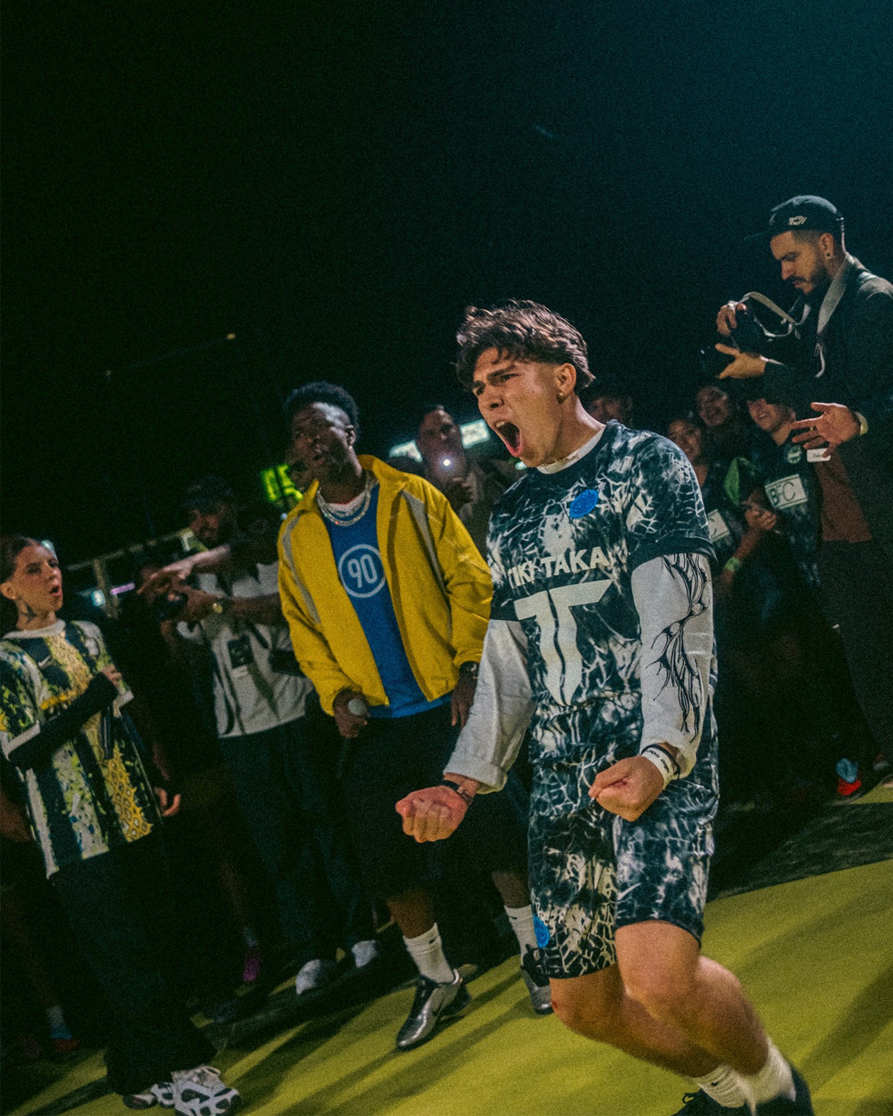 A young man in sportswear celebrates energetically on an indoor field, surrounded by people watching and taking photos.