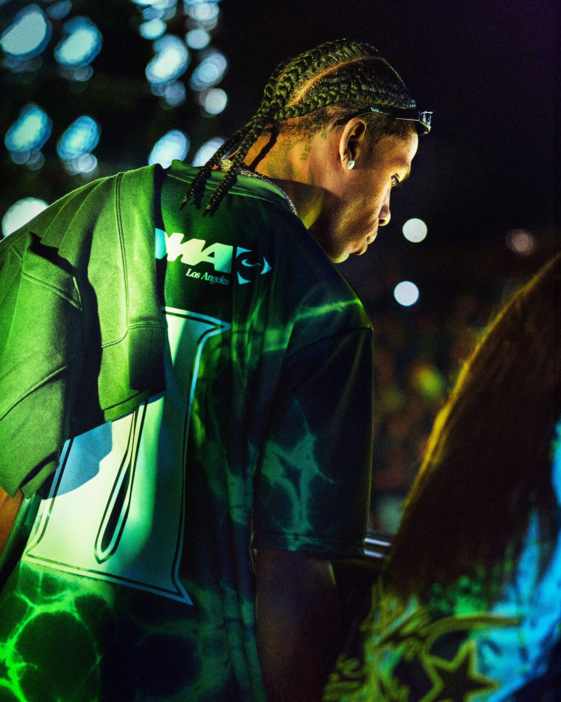 A man with braided hair wearing a green sports jersey stands under colorful lights, facing away from the camera at a nighttime event.