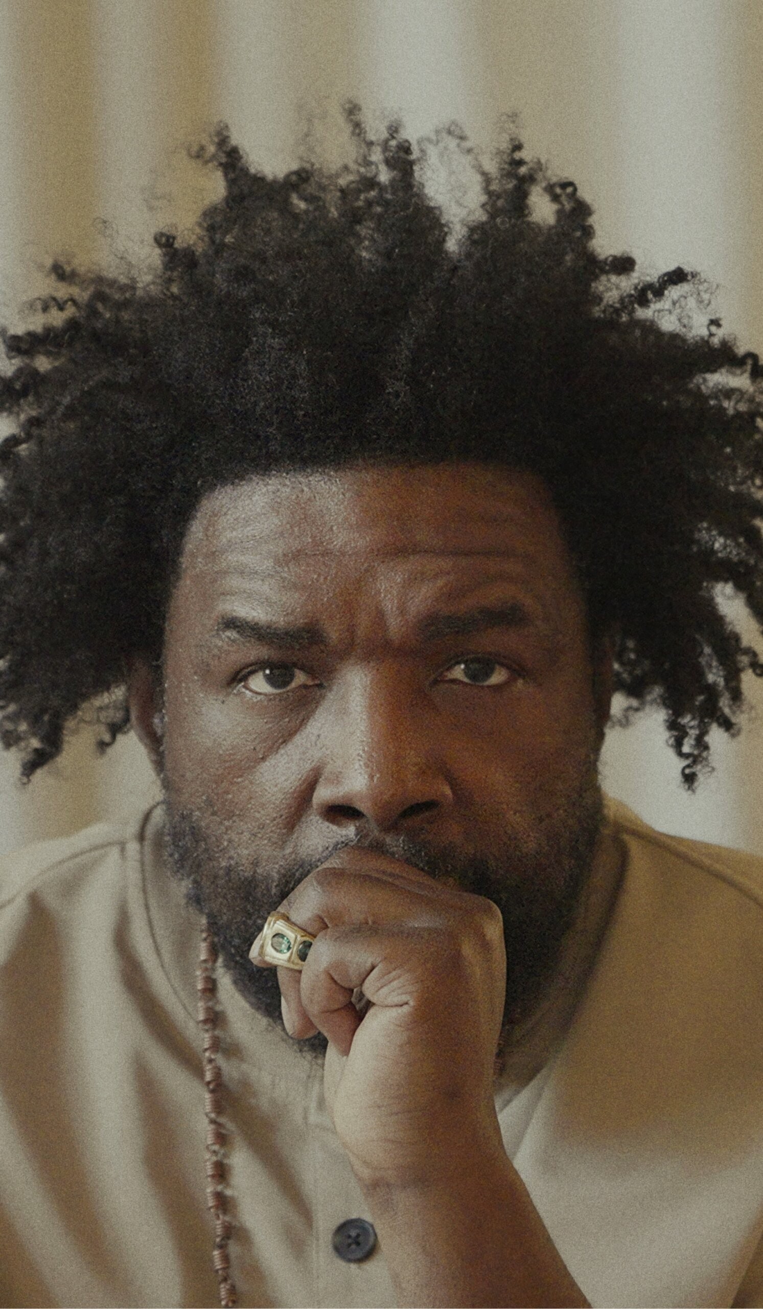 A man with a beard and natural curly hair rests his chin on his hand, looking directly at the camera. He wears a beige shirt, a beaded necklace, and a ring.