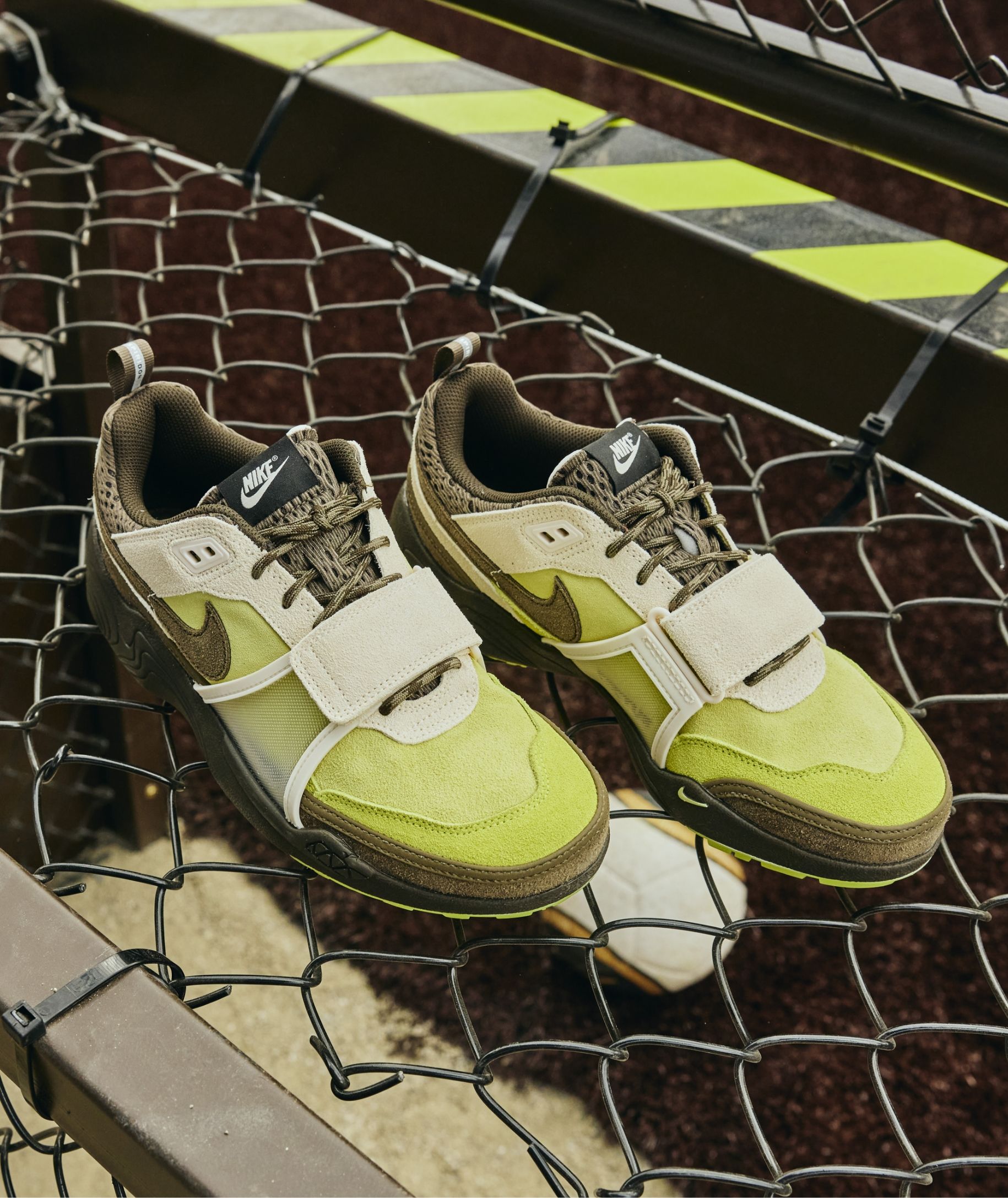 A pair of green, brown, and white Nike sneakers with velcro straps placed on a metal fence with yellow and black caution tape.