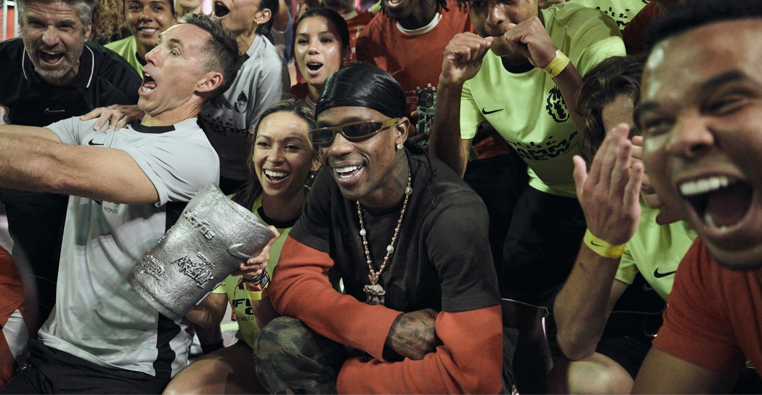 A group of people, some wearing sportswear, gather for a lively group photo. One person, Travis Scott, at the center wears sunglasses and a headband.