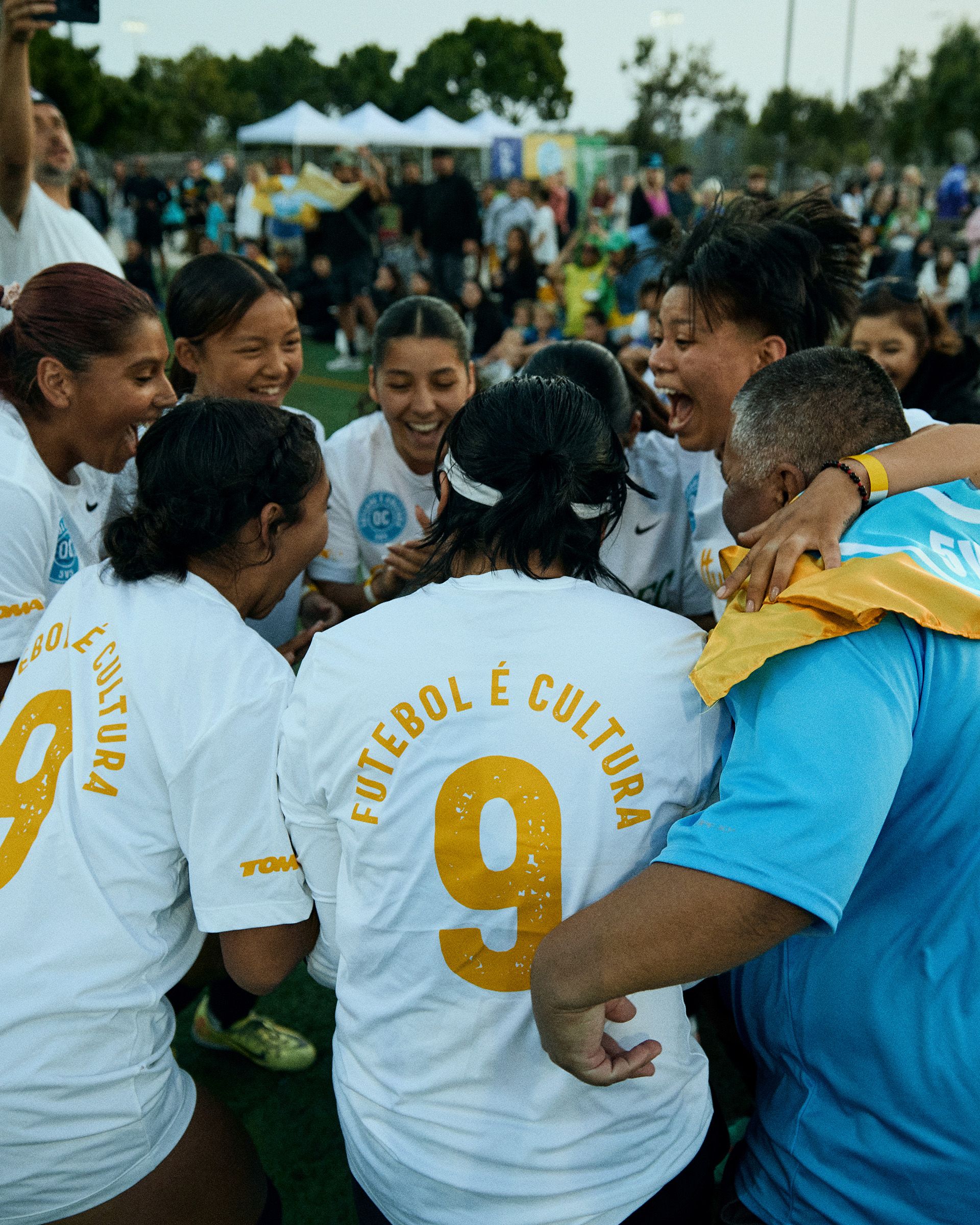 A group of soccer players wearing white jerseys with "Futebol É Cultura" on the back stand in a circle, celebrating together on a field with a crowd in the background.