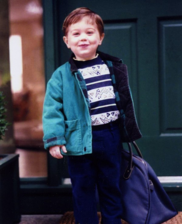 A young child stands in front of a green door, wearing a teal jacket, patterned shirt, navy pants, and colorful shoes, holding a blue bag.