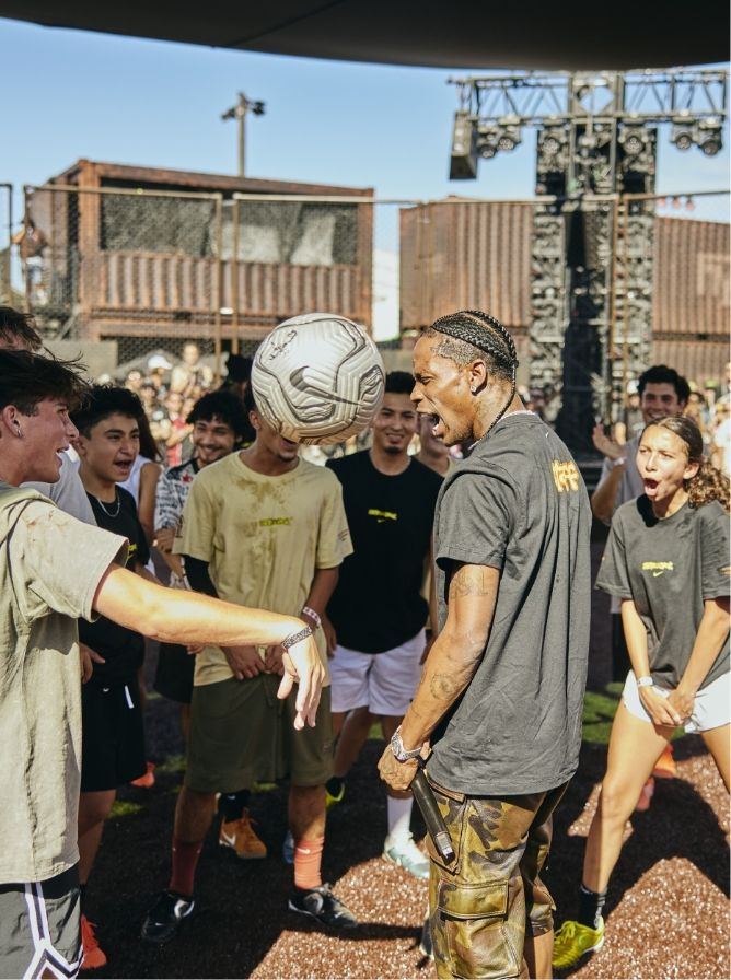 A group of people stand outdoors in a circle, reacting as a soccer ball is about to hit a man in the face. Some are laughing and others are surprised.