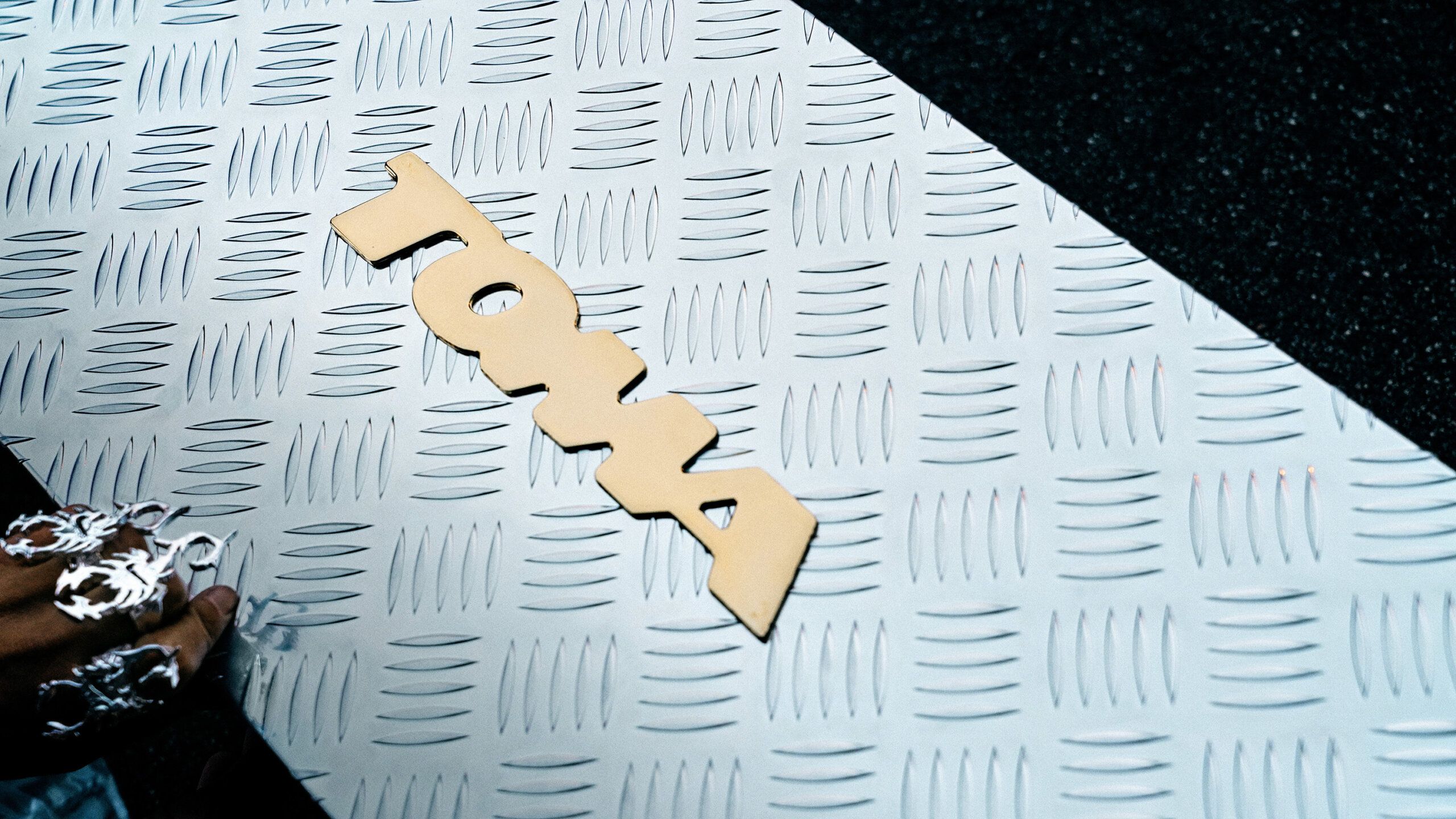 A cutout of the word "tow" lies on a textured metal surface, with part of a hand and decorative elements visible in the bottom left corner.