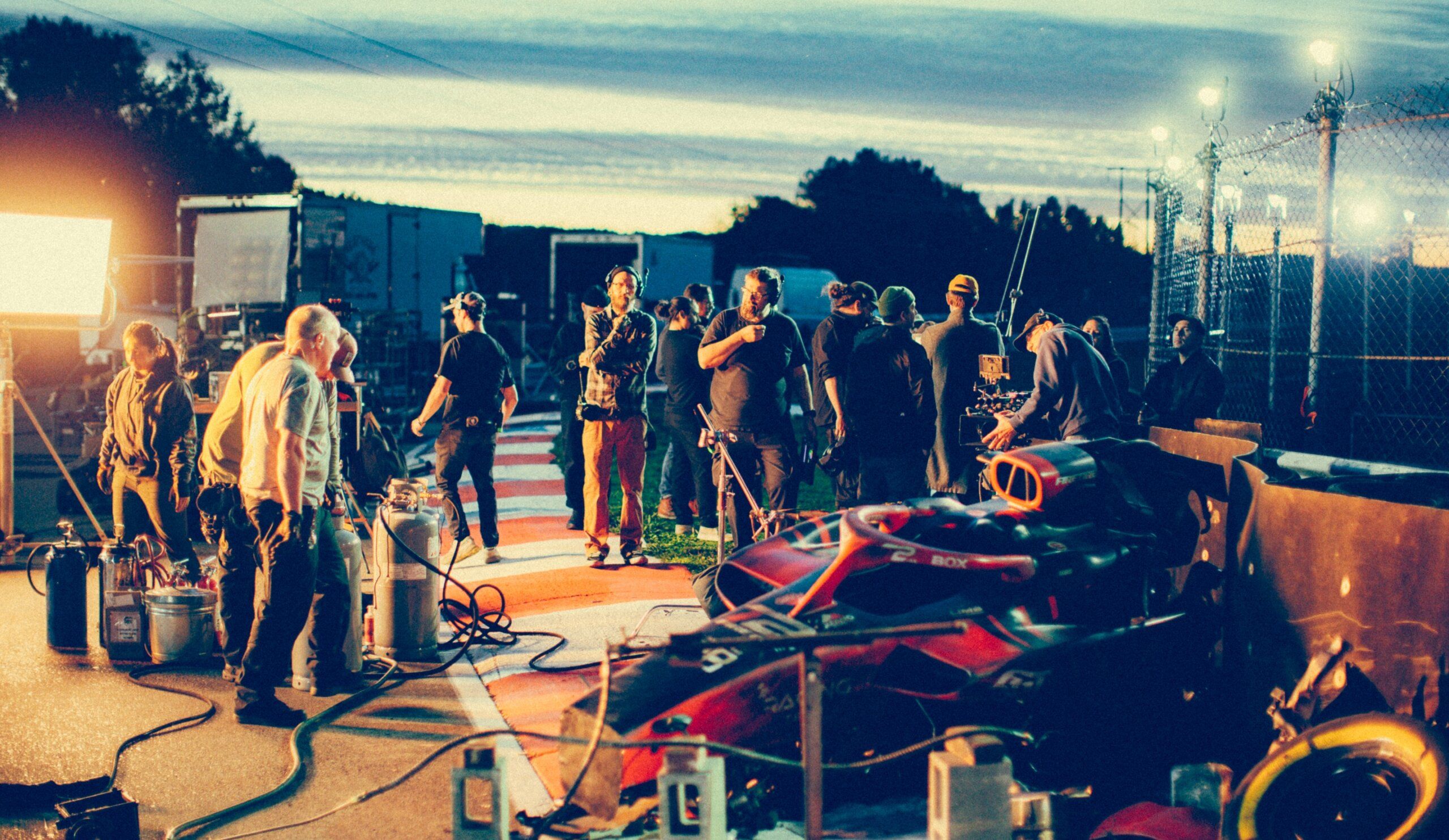 Film crew working outdoors during dusk, with lighting equipment and a racing car visible.