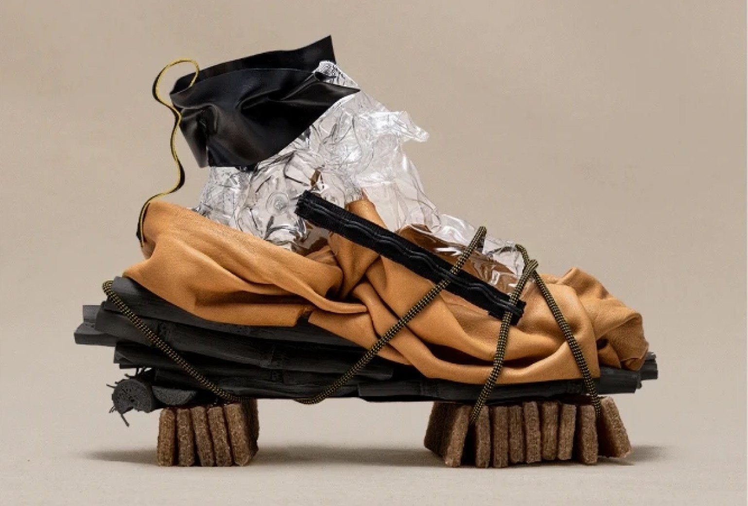 An abstract sculpture resembling a shoe, made of layered materials including black fabric, clear plastic, tan cloth, cord, and sponge-like blocks, displayed against a plain background.