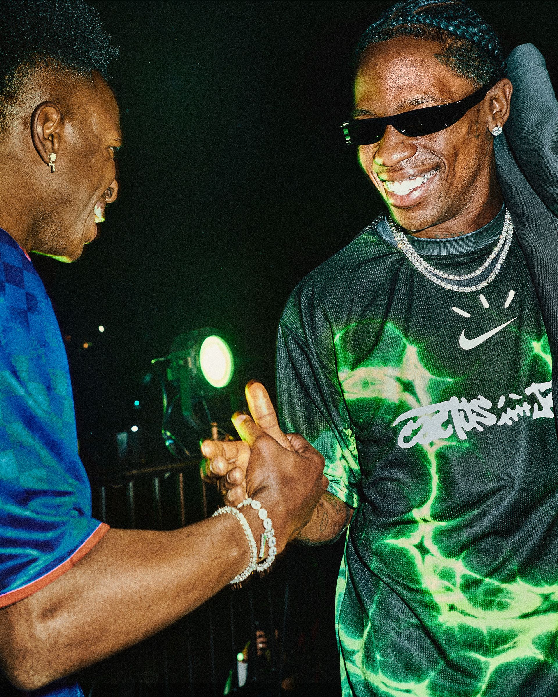 Two men greet each other with a handshake at a nighttime event, both smiling; one wears dark sunglasses and a green patterned shirt, the other wears a blue shirt.