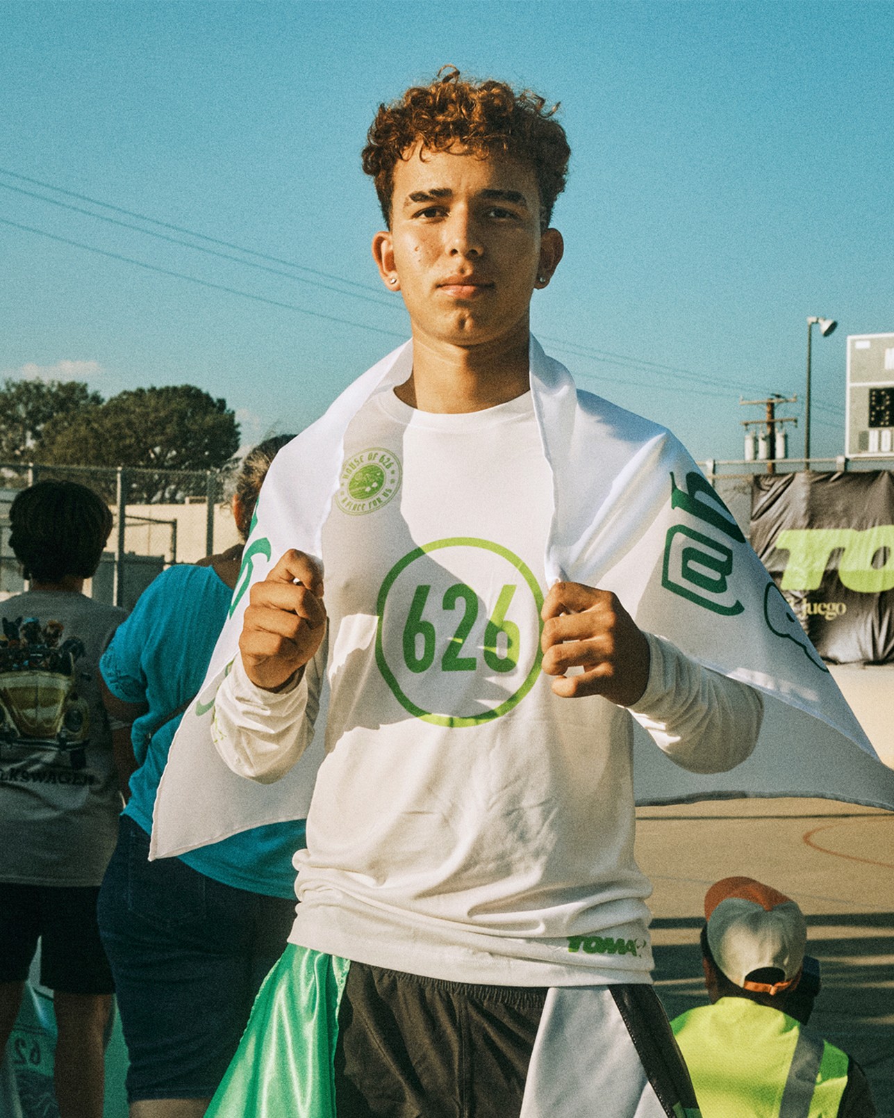 A young person wearing a white “626” shirt and holding a matching flag stands at an outdoor event, with other people and a scoreboard visible in the background.