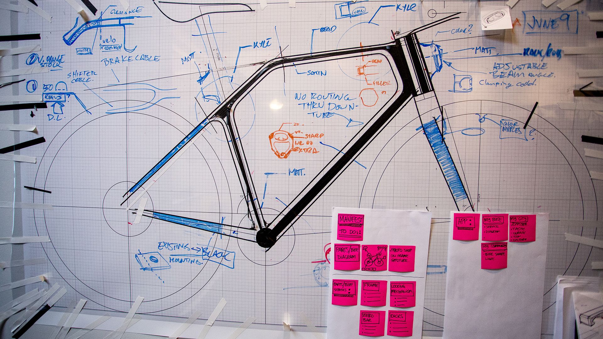 Bicycle frame prototype hanging on a wall covered in design notes and diagrams.