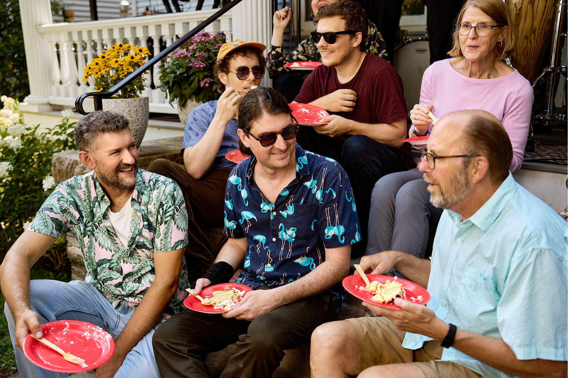Six adults sit on outdoor steps eating food from red plates, wearing casual summer clothing, and smiling as they engage in conversation.