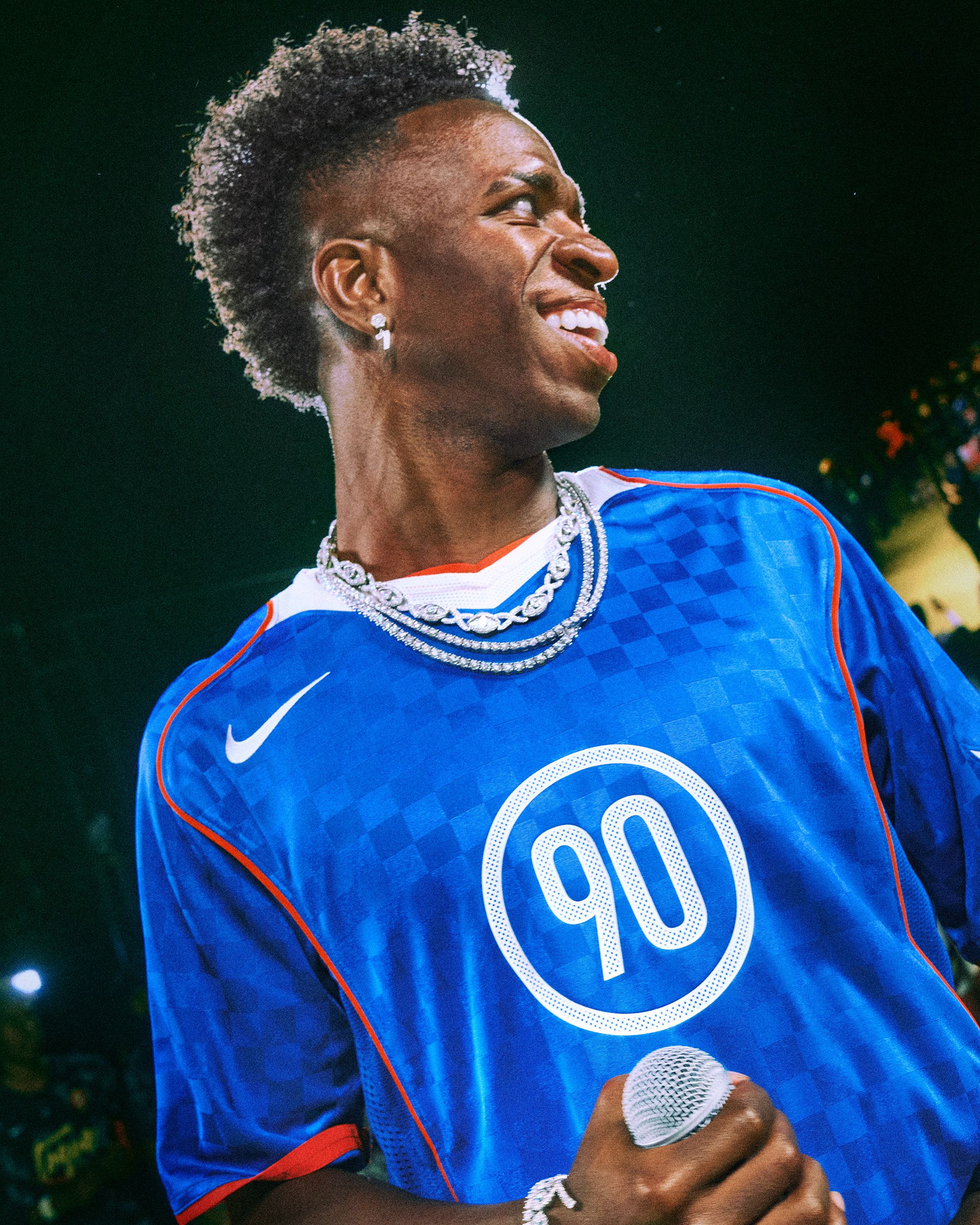 A person in a blue sports jersey with the number 90 holds a microphone and smiles, wearing layered silver necklaces under stage lighting.