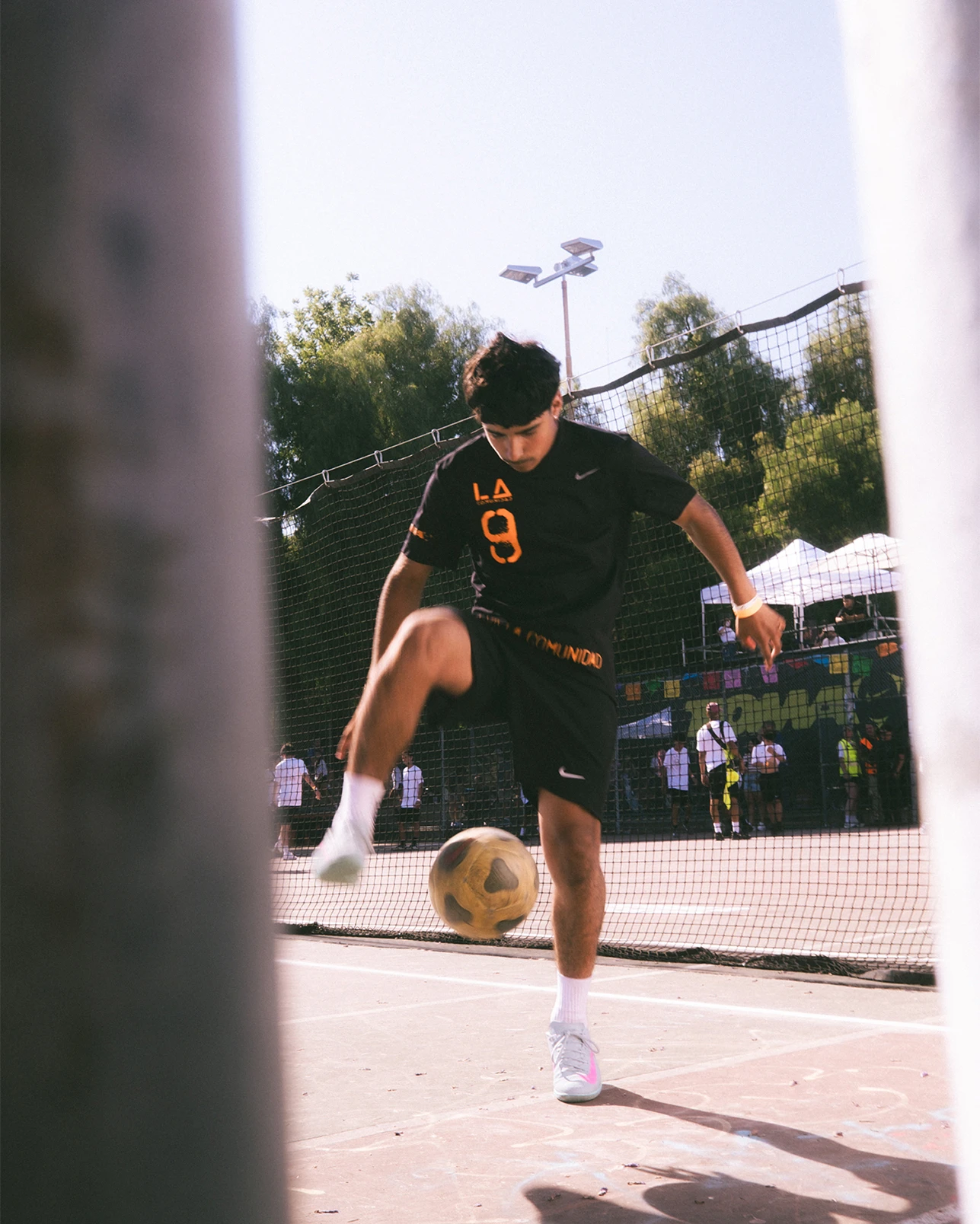 A person in a black sports uniform balances a soccer ball on one foot on an outdoor court, with a net and trees in the background.