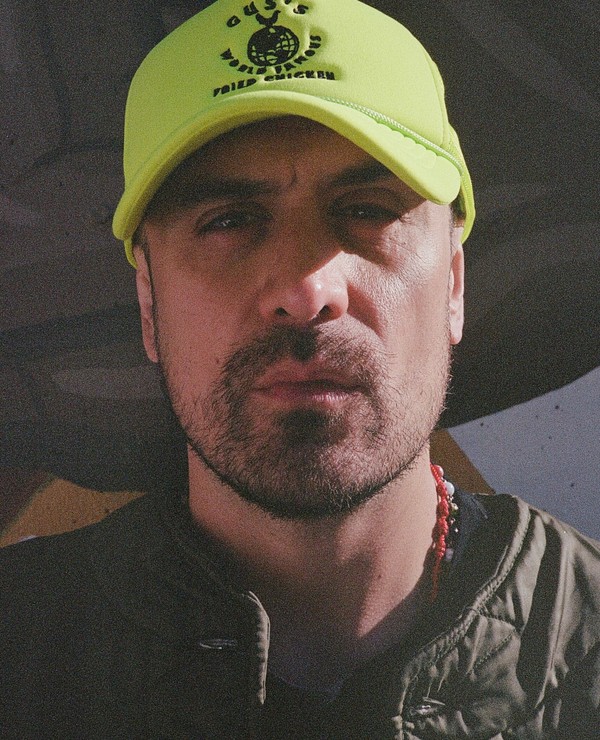 A man with a beard and mustache wears a bright yellow cap and a green jacket, standing in front of a blurred background.