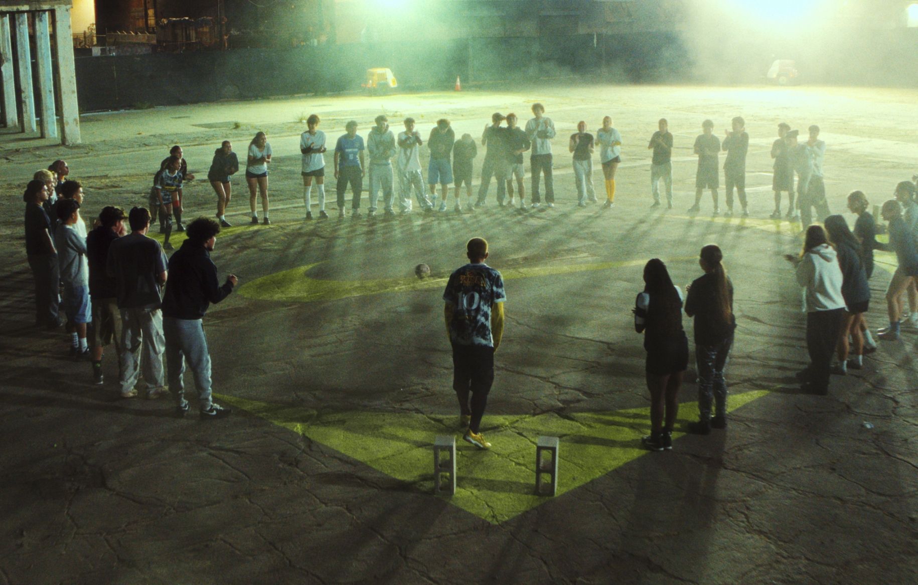 A group of people stand in a large circle at night on a cracked concrete surface, illuminated by artificial lights, with a person in the center.