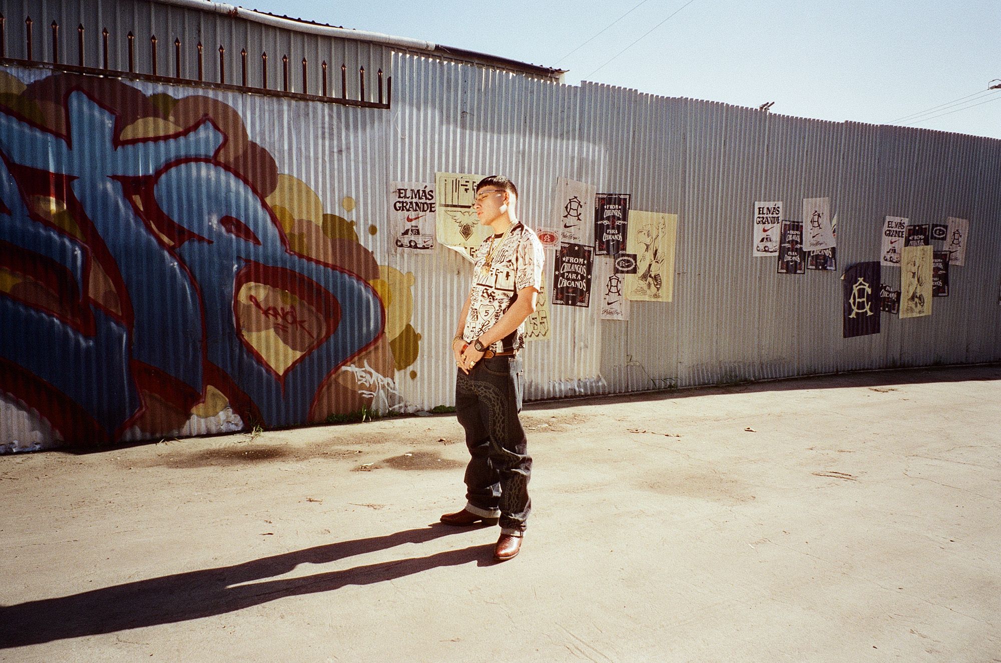 A person stands outside on a sunny day near a corrugated metal fence with graffiti and posters. The ground casts a long shadow.