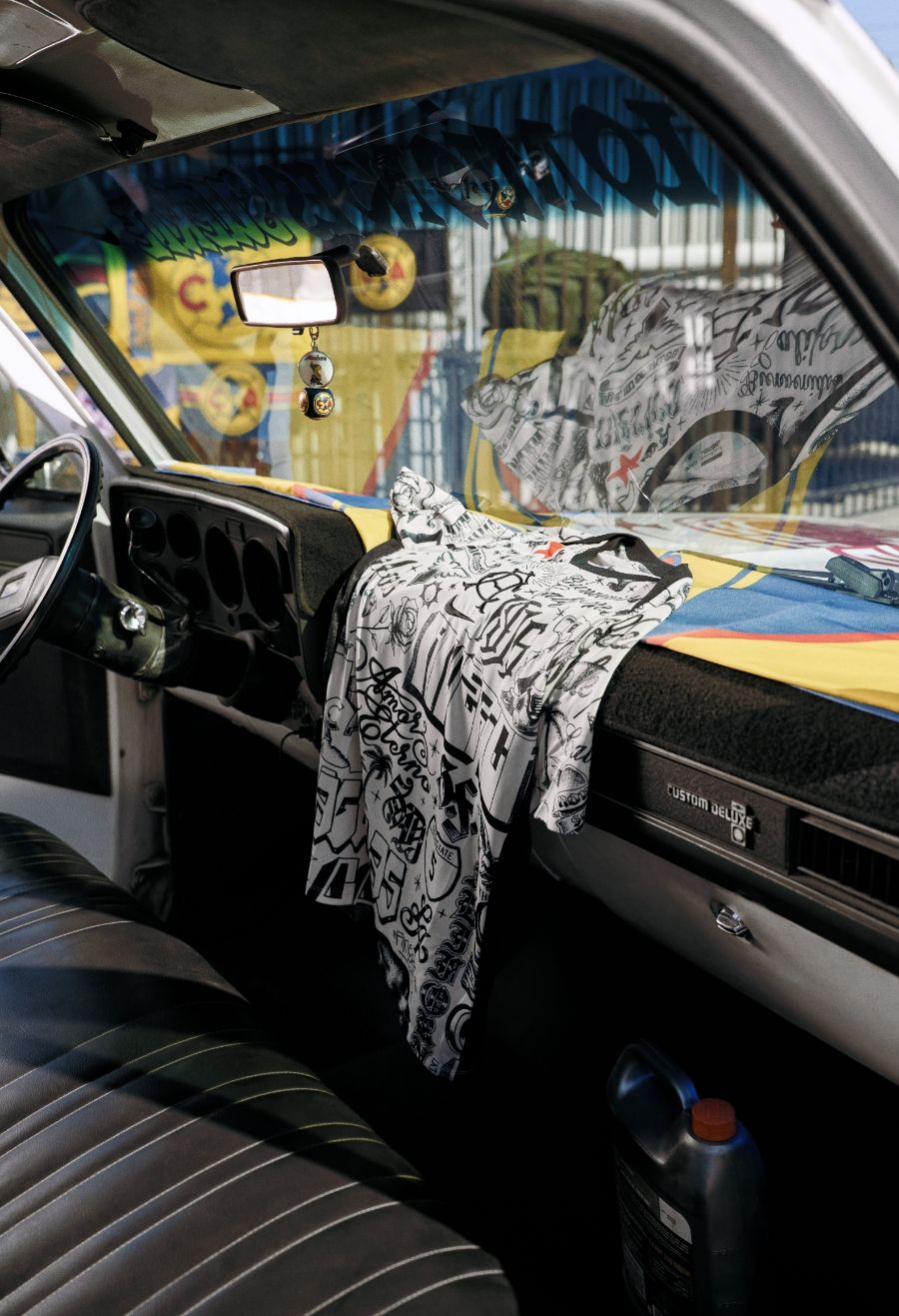 A graphic t-shirt with black and white designs is draped over the dashboard of a vintage car with a colorful windshield visor and black vinyl seats.