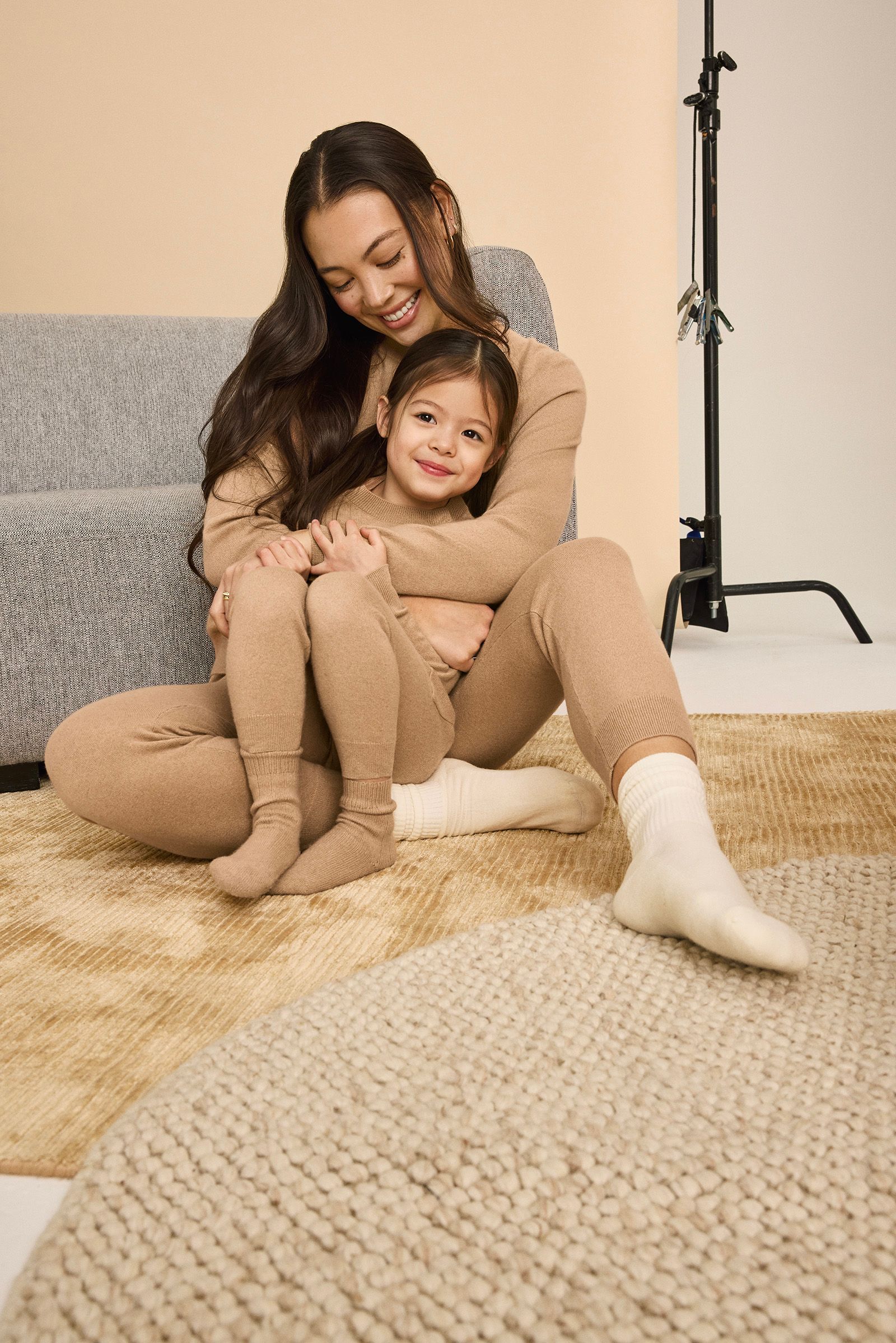 A woman and young girl, both dressed in matching tan loungewear, sit on a beige rug in a cozy room, smiling and embracing near a gray sofa.
